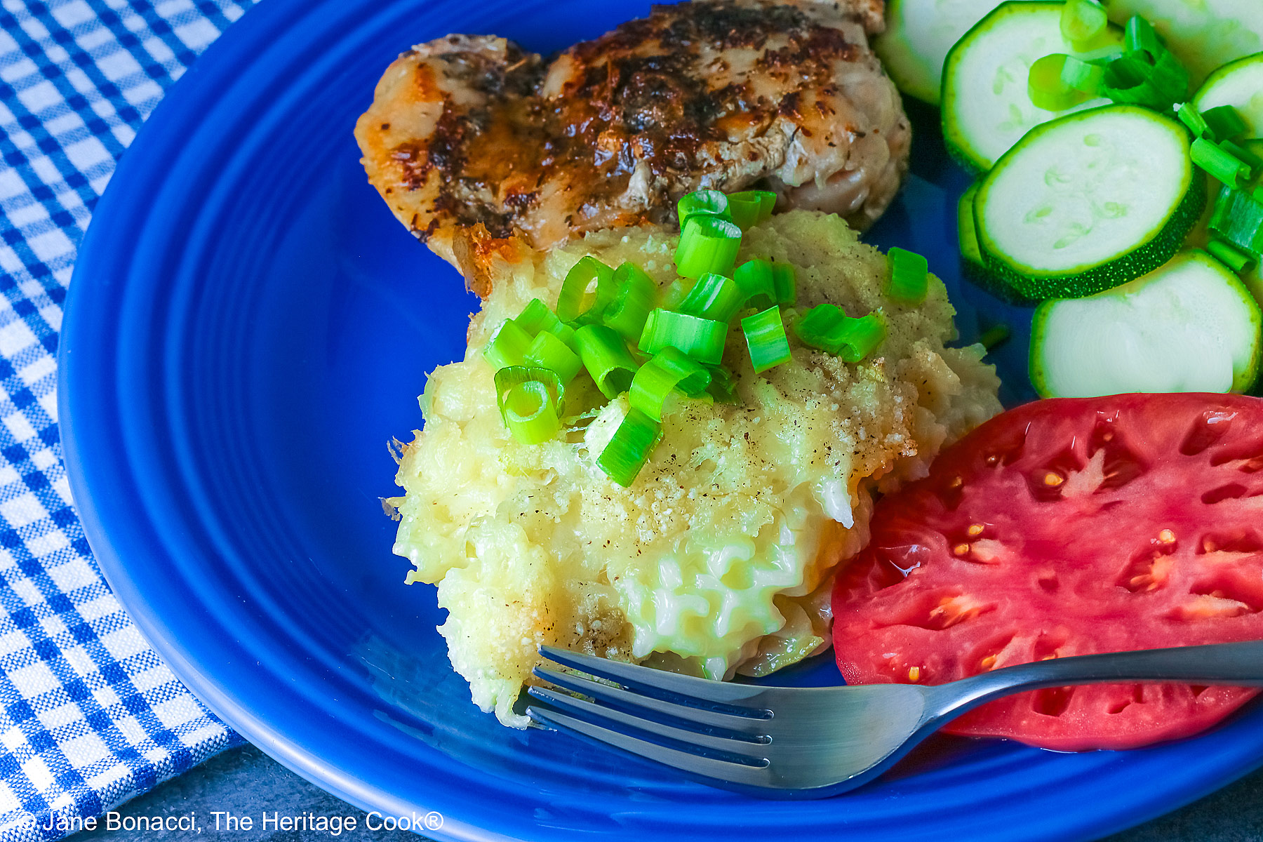 Bright blue plate filled with a piece of chicken, sliced zucchini, an heirloom tomato, large scoop of Cheesy Chile Rice Casserole topped with sliced green onions, with a blue checked cloth in the back and a fork in front © 2026 Jane Bonacci, The Heritage Cook.
