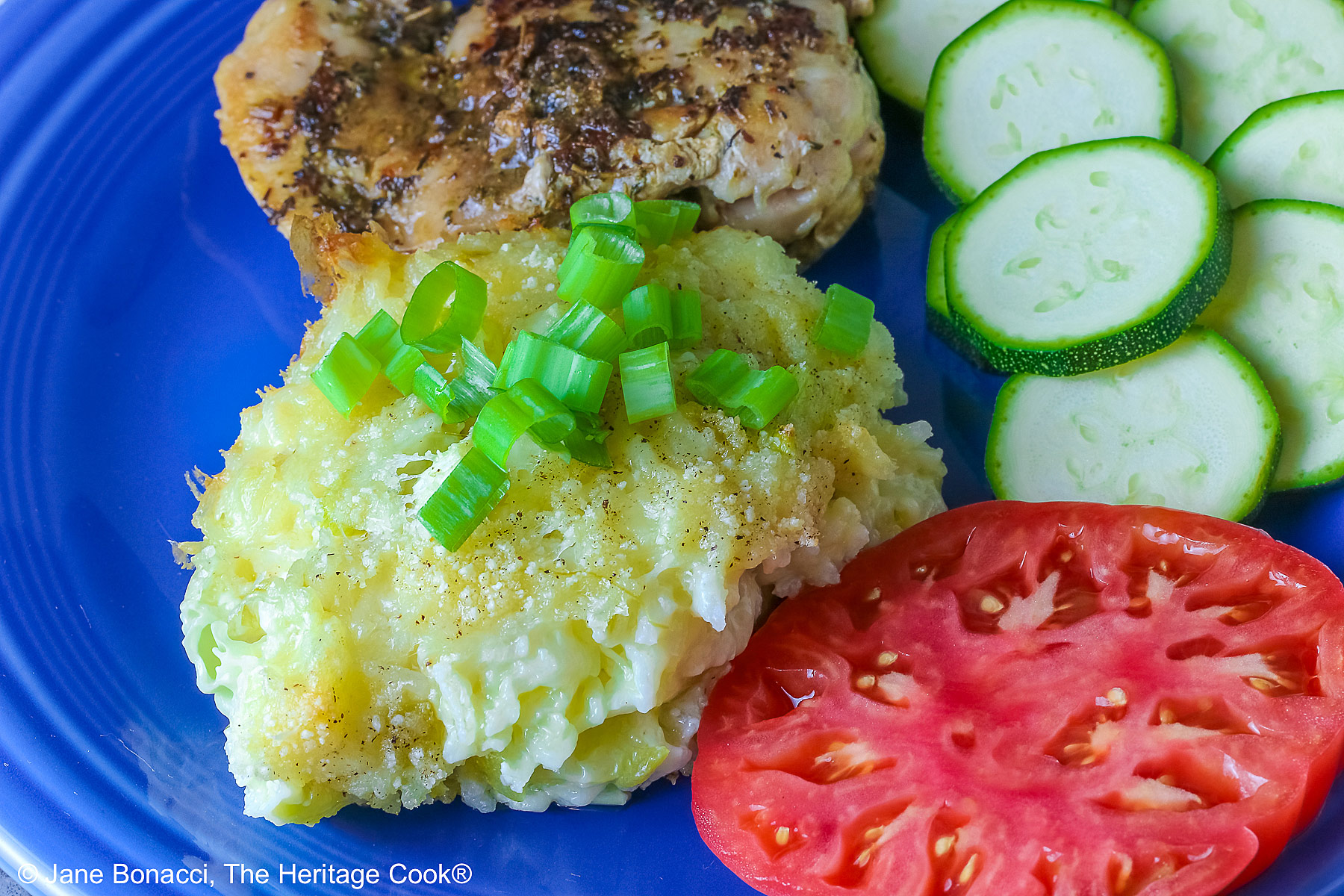 Bright blue plate filled with a piece of chicken, sliced zucchini, an heirloom tomato, large scoop of Cheesy Chile Rice Casserole topped with sliced green onions, with a blue checked cloth in the back and a fork in front © 2026 Jane Bonacci, The Heritage Cook.