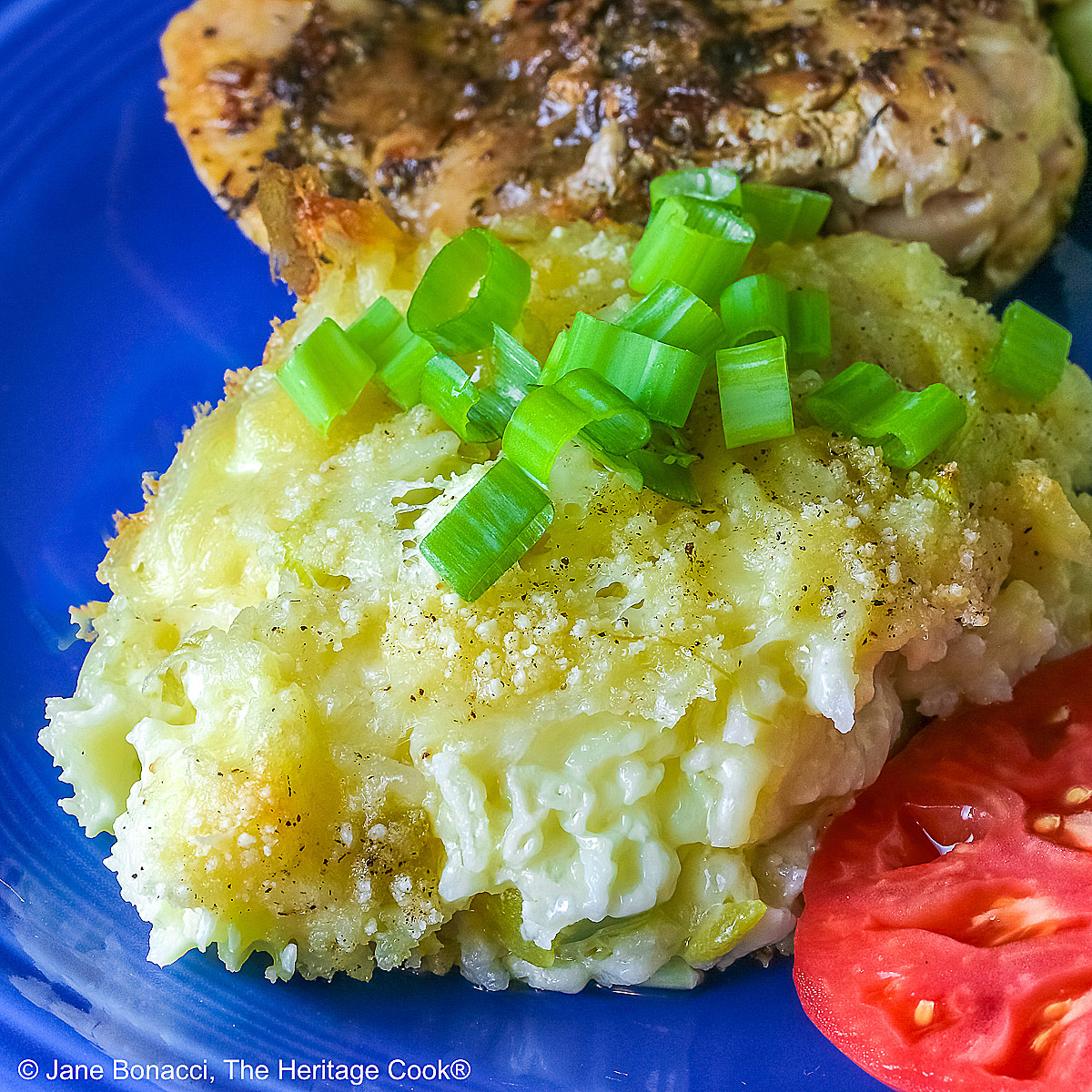 Close shot of rice; Bright blue plate filled with a piece of chicken, sliced zucchini, an heirloom tomato, large scoop of Cheesy Chile Rice Casserole topped with sliced green onions, with a blue checked cloth in the back and a fork in front © 2026 Jane Bonacci, The Heritage Cook.