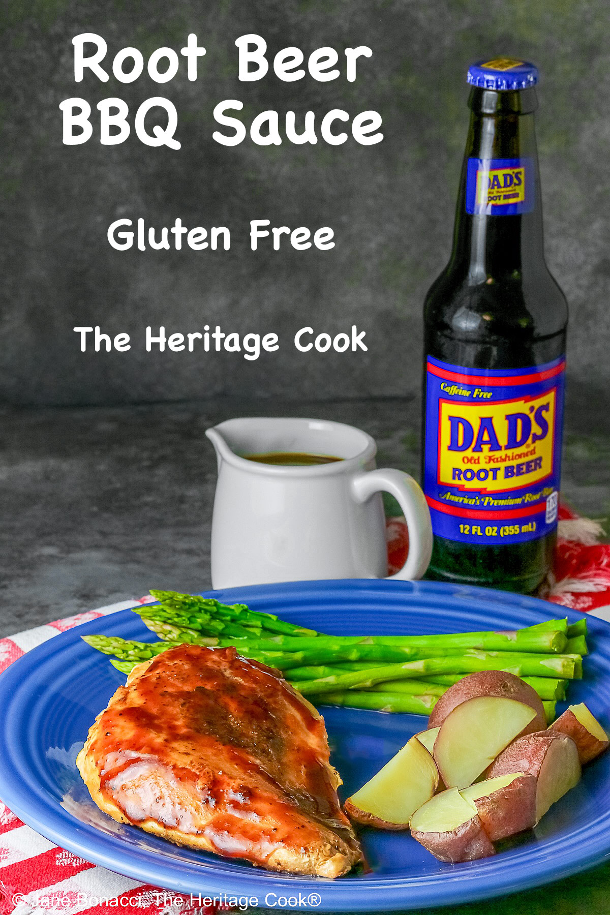 A chicken breast coated with Root Beer BBQ Sauce on a blue plate with asparagus and red potatoes with a small white pitcher of the BBQ sauce and a bottle of root beer behind the plate sitting on a red and white checked cloth © 2026 Jane Bonacci, The Heritage Cook.