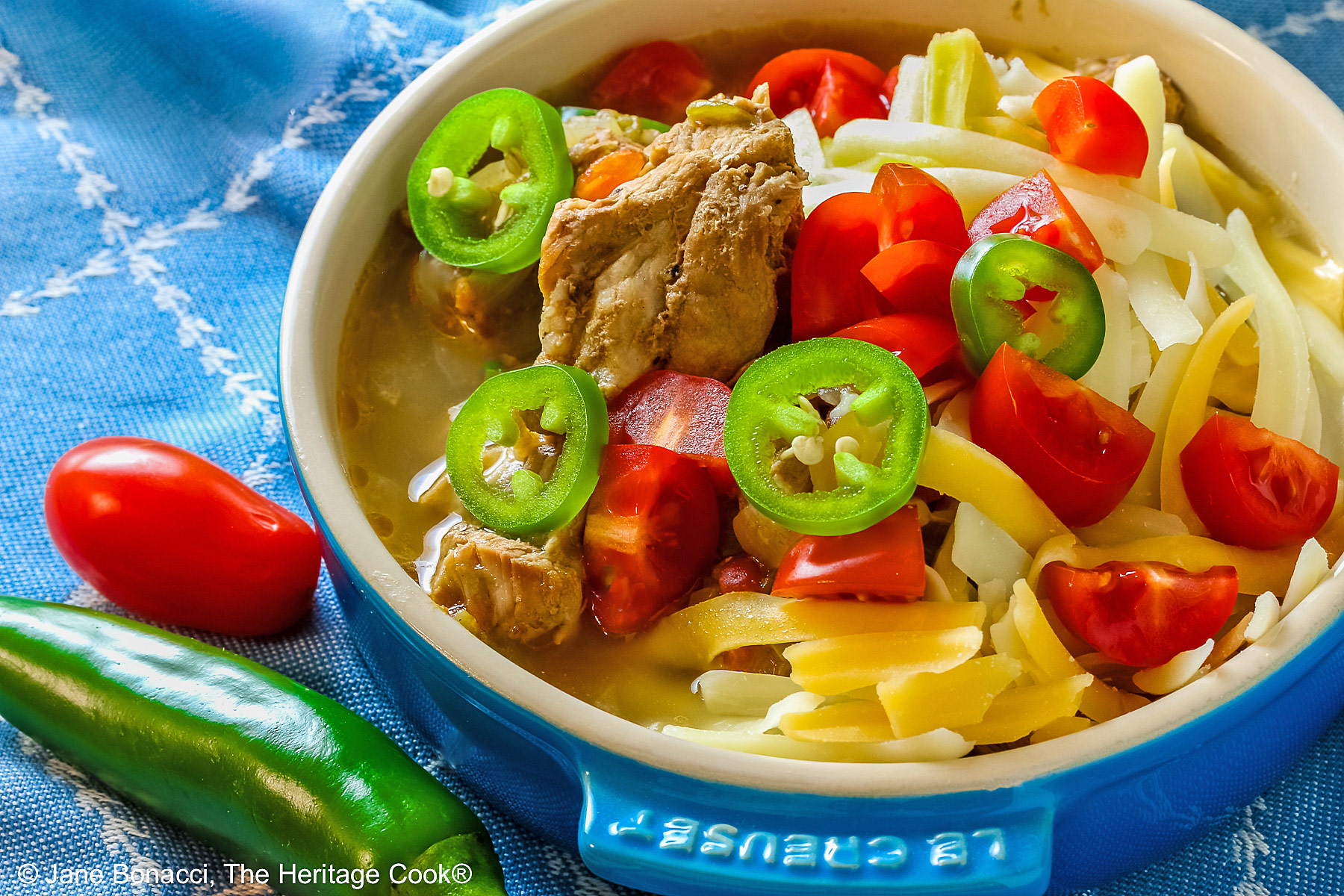 Blue bowl brimming with Spicy Cheesy Chile Pork Stew topped with sliced jalapenos, shredded cheese, and chopped tomatoes sitting on a decorative blue cloth © 2026 Jane Bonacci, The Heritage Cook.