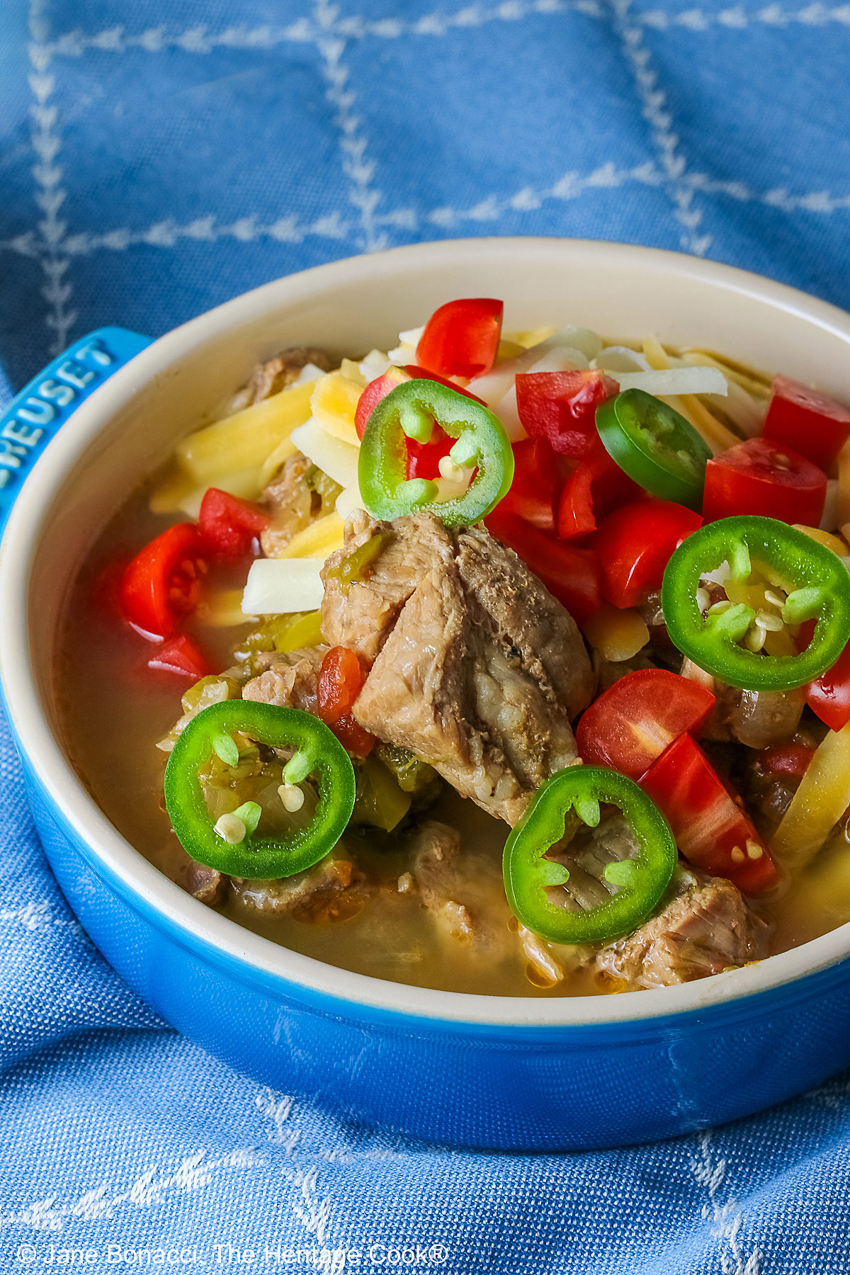 Blue bowl brimming with Spicy Cheesy Chile Pork Stew topped with sliced jalapenos, shredded cheese, and chopped tomatoes sitting on a decorative blue cloth © 2026 Jane Bonacci, The Heritage Cook.