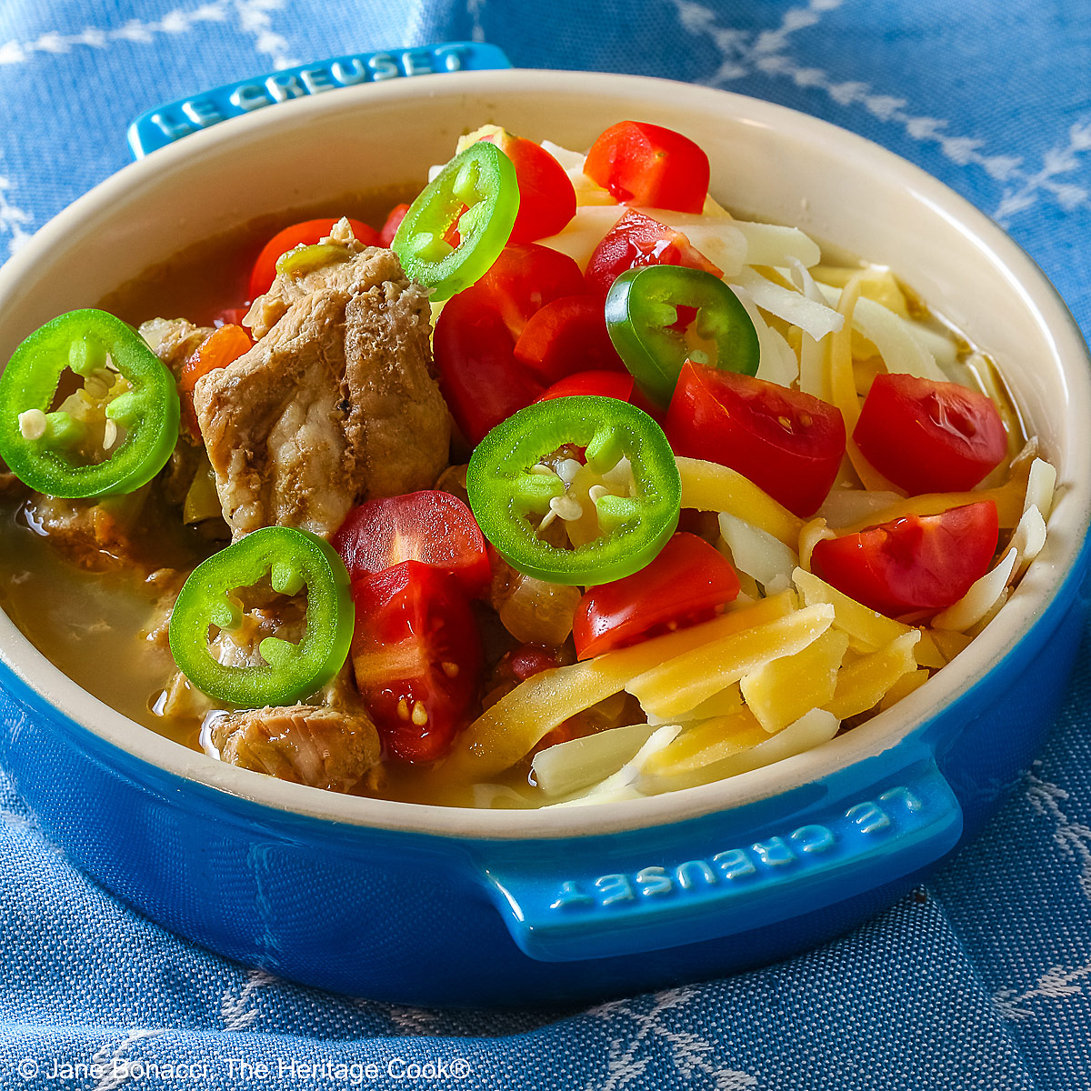 Blue bowl brimming with Spicy Cheesy Chile Pork Stew topped with sliced jalapenos, shredded cheese, and chopped tomatoes sitting on a decorative blue cloth © 2026 Jane Bonacci, The Heritage Cook.
