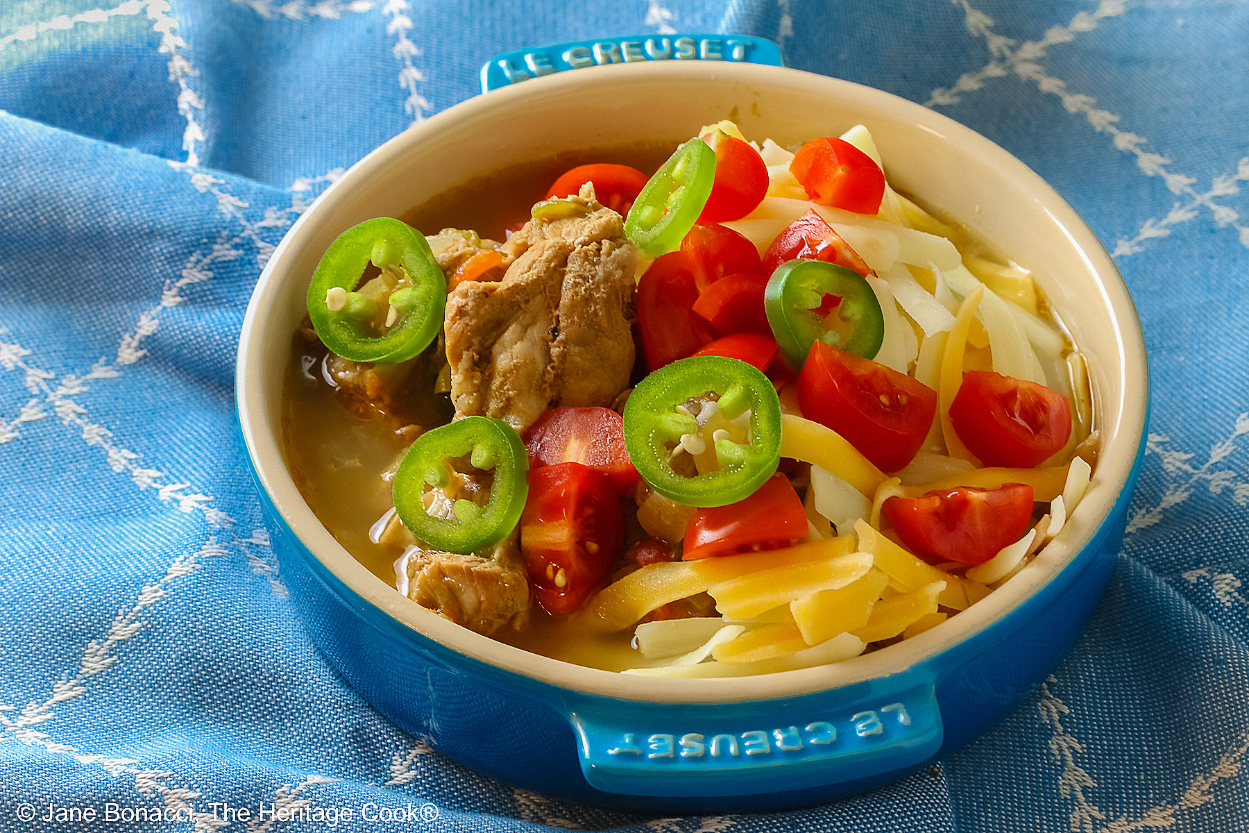 Blue bowl brimming with Spicy Cheesy Chile Pork Stew topped with sliced jalapenos, shredded cheese, and chopped tomatoes sitting on a decorative blue cloth © 2026 Jane Bonacci, The Heritage Cook.