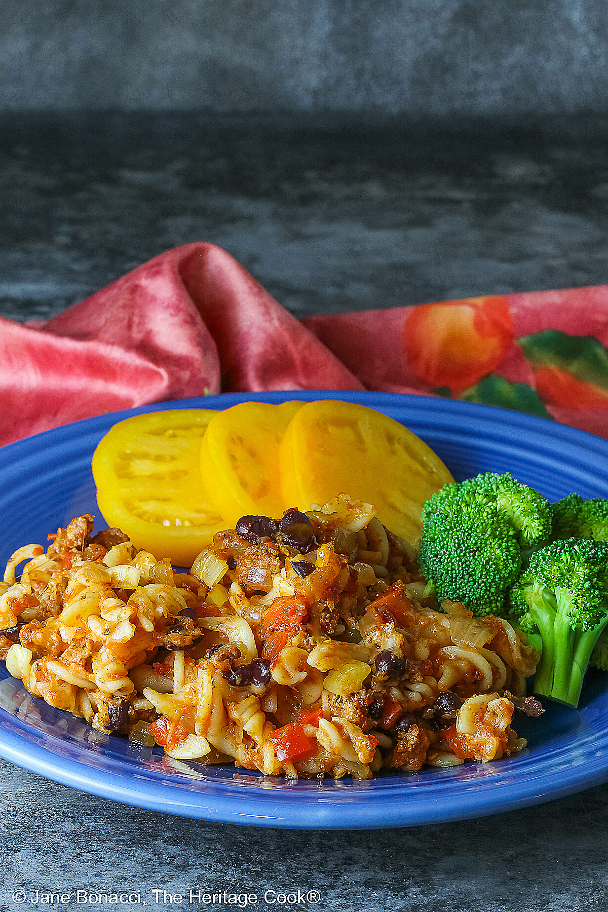 Large scoop of Cheesy Taco Pasta Casserole on a dark blue plate flanked by broccoli and sliced yellow tomatoes with a deep magenta cloth in the back on a dark gray background © 2025 Jane Bonacci, The Heritage Cook.