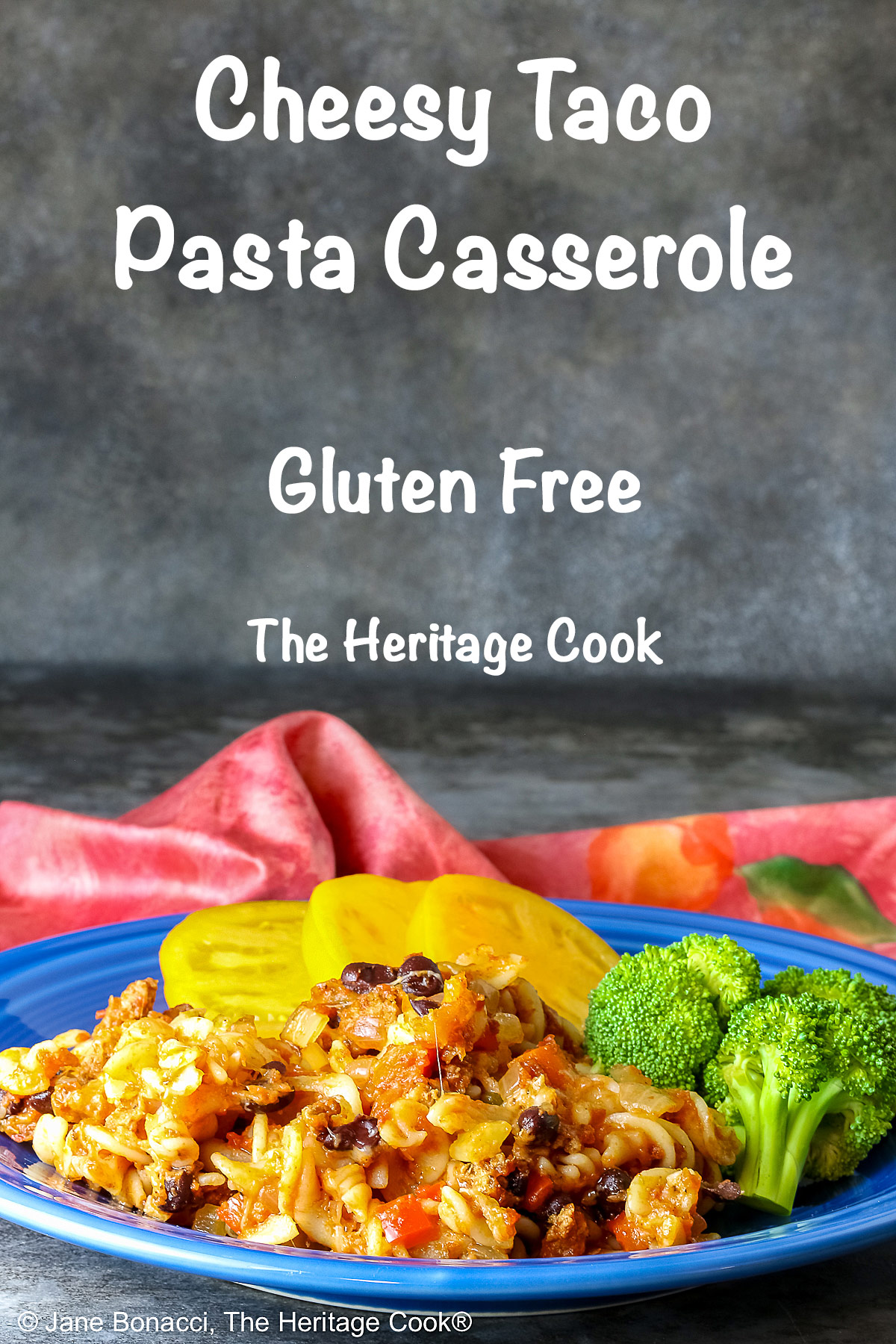 Large scoop of Cheesy Taco Pasta Casserole on a dark blue plate flanked by broccoli and sliced yellow tomatoes with a deep magenta cloth in the back on a dark gray background © 2025 Jane Bonacci, The Heritage Cook.