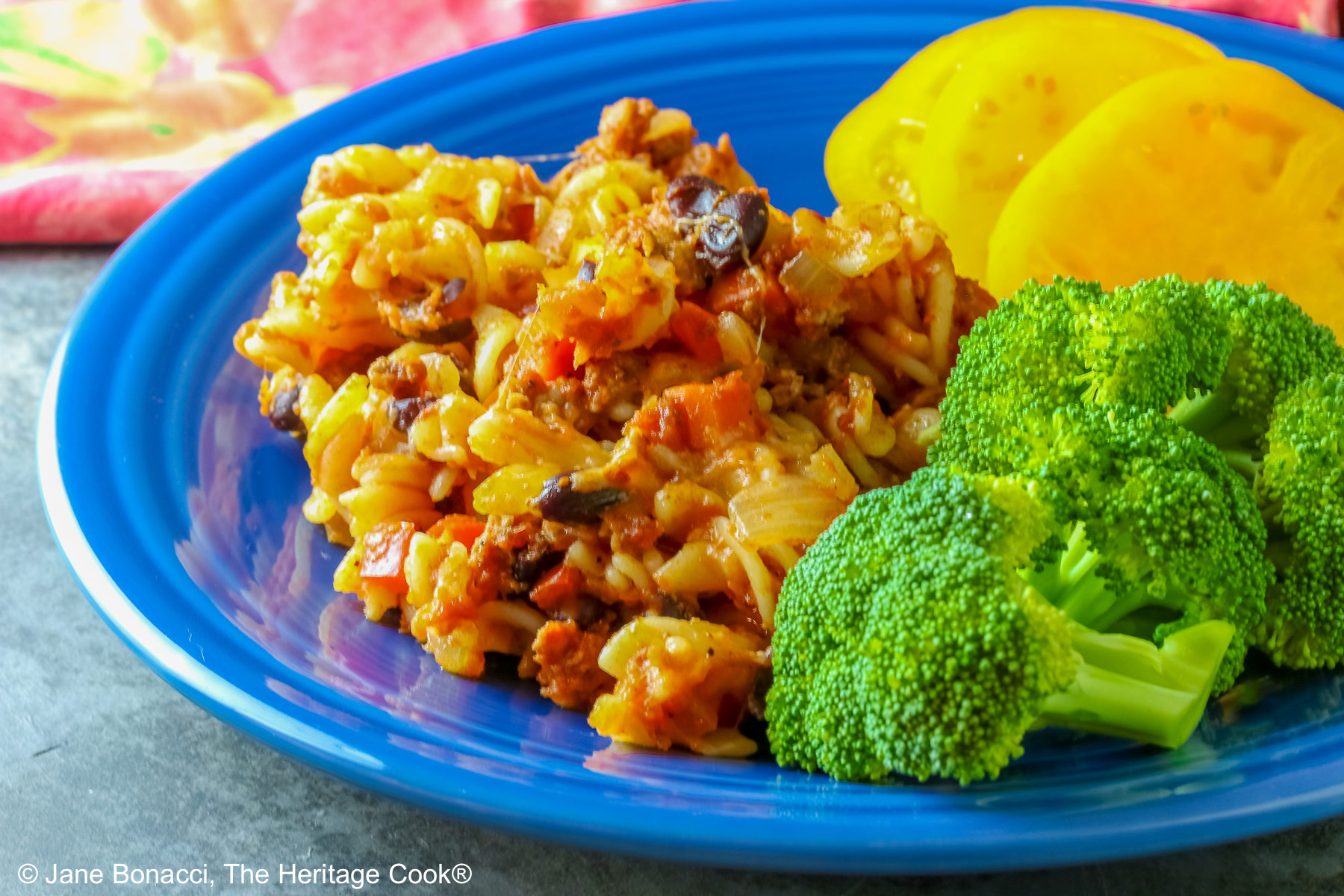 Large scoop of Cheesy Taco Pasta Casserole on a dark blue plate flanked by broccoli and sliced yellow tomatoes with a deep magenta cloth in the back on a dark gray background © 2025 Jane Bonacci, The Heritage Cook.