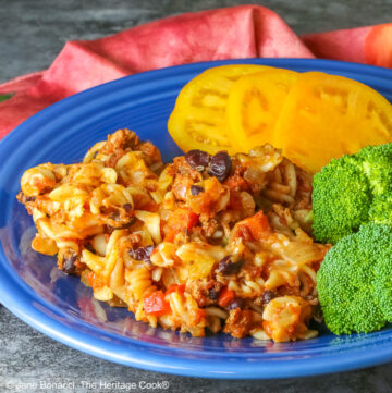 Large scoop of Cheesy Taco Pasta Casserole on a dark blue plate flanked by broccoli and sliced yellow tomatoes with a deep magenta cloth in the back on a dark gray background © 2025 Jane Bonacci, The Heritage Cook.