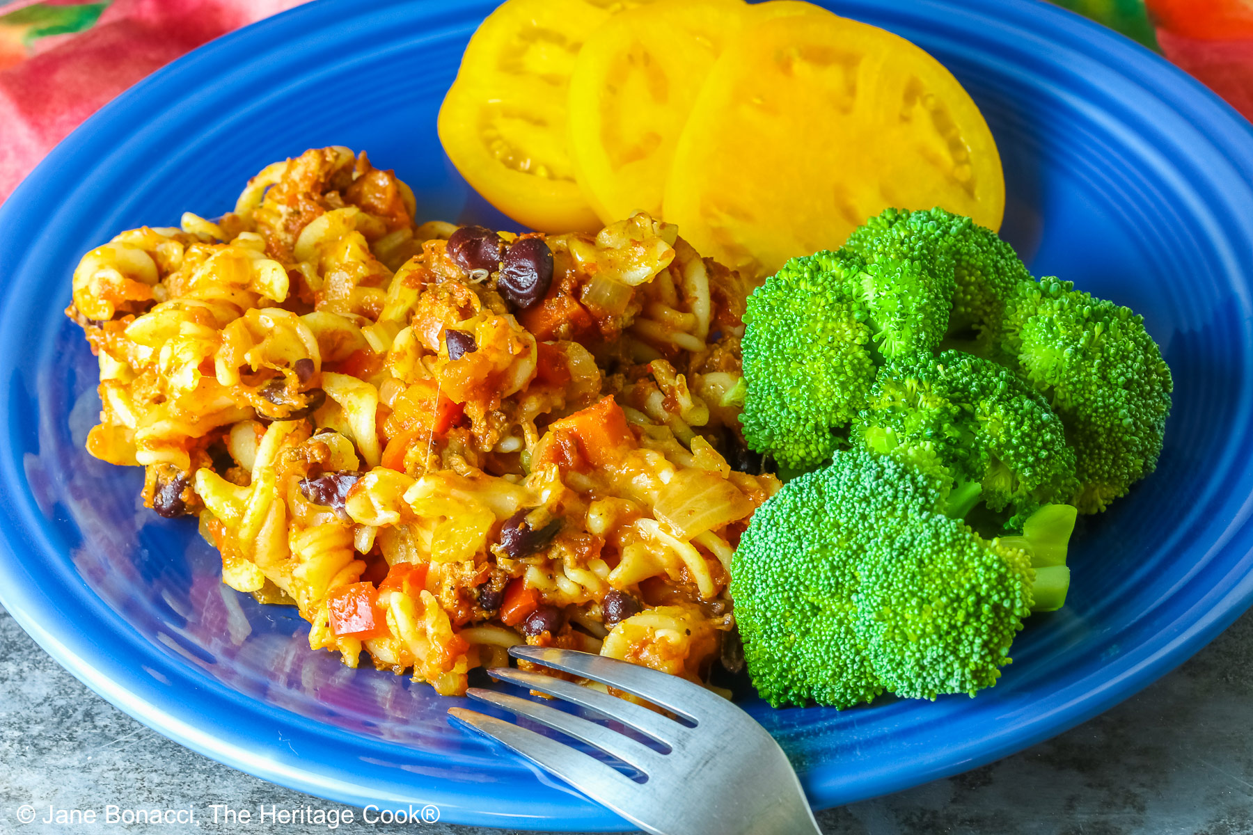 Large scoop of Cheesy Taco Pasta Casserole on a dark blue plate flanked by broccoli and sliced yellow tomatoes with a deep magenta cloth in the back on a dark gray background © 2025 Jane Bonacci, The Heritage Cook.