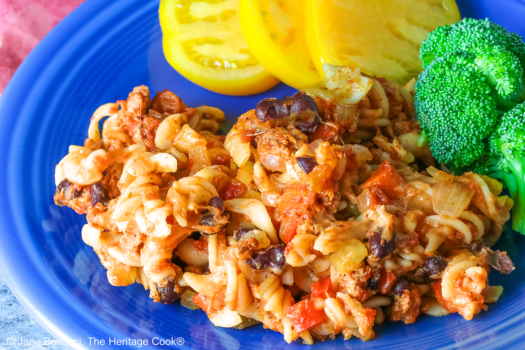 Large scoop of Cheesy Taco Pasta Casserole on a dark blue plate flanked by broccoli and sliced yellow tomatoes with a deep magenta cloth in the back on a dark gray background © 2025 Jane Bonacci, The Heritage Cook.