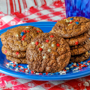 Two short stacks of decorated Intense Chocolate Mudslide Cookies on a vivid blue plate with a single cookie propped up in front of the stacks, sitting on red and white cloths with small blue pitcher behind; the cookies and plate are decorated with red, white, and blue candy stars for Memorial Day © 2025 Jane Bonacci, The Heritage Cook.