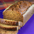 Slices in front of a loaf on purple cutting board with an orange plaid cloth beneath; Harvest Pumpkin Bread © 2022 Jane Bonacci, The Heritage Cook.
