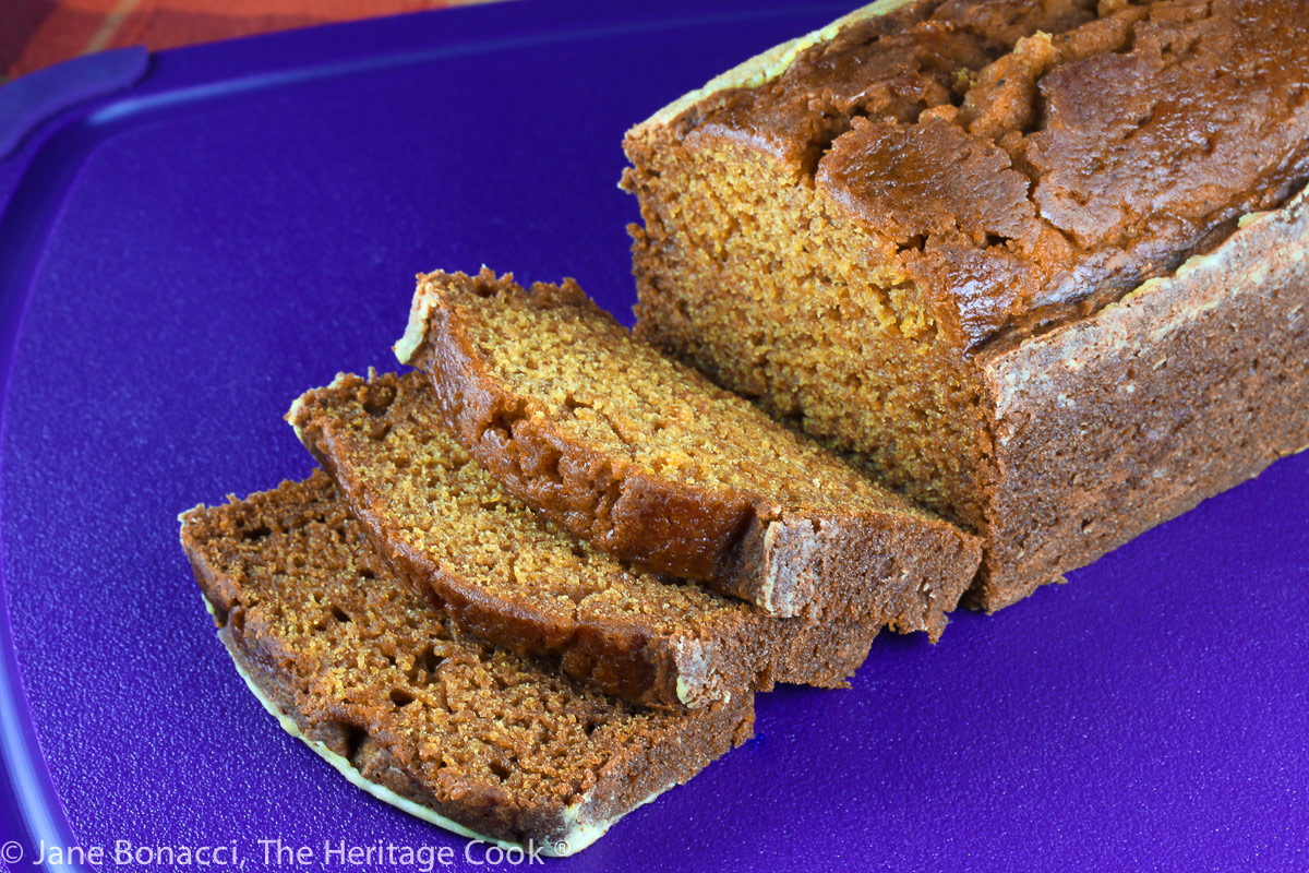 Slices in front of a loaf on purple cutting board with an orange plaid cloth beneath; Harvest Pumpkin Bread © 2022 Jane Bonacci, The Heritage Cook.