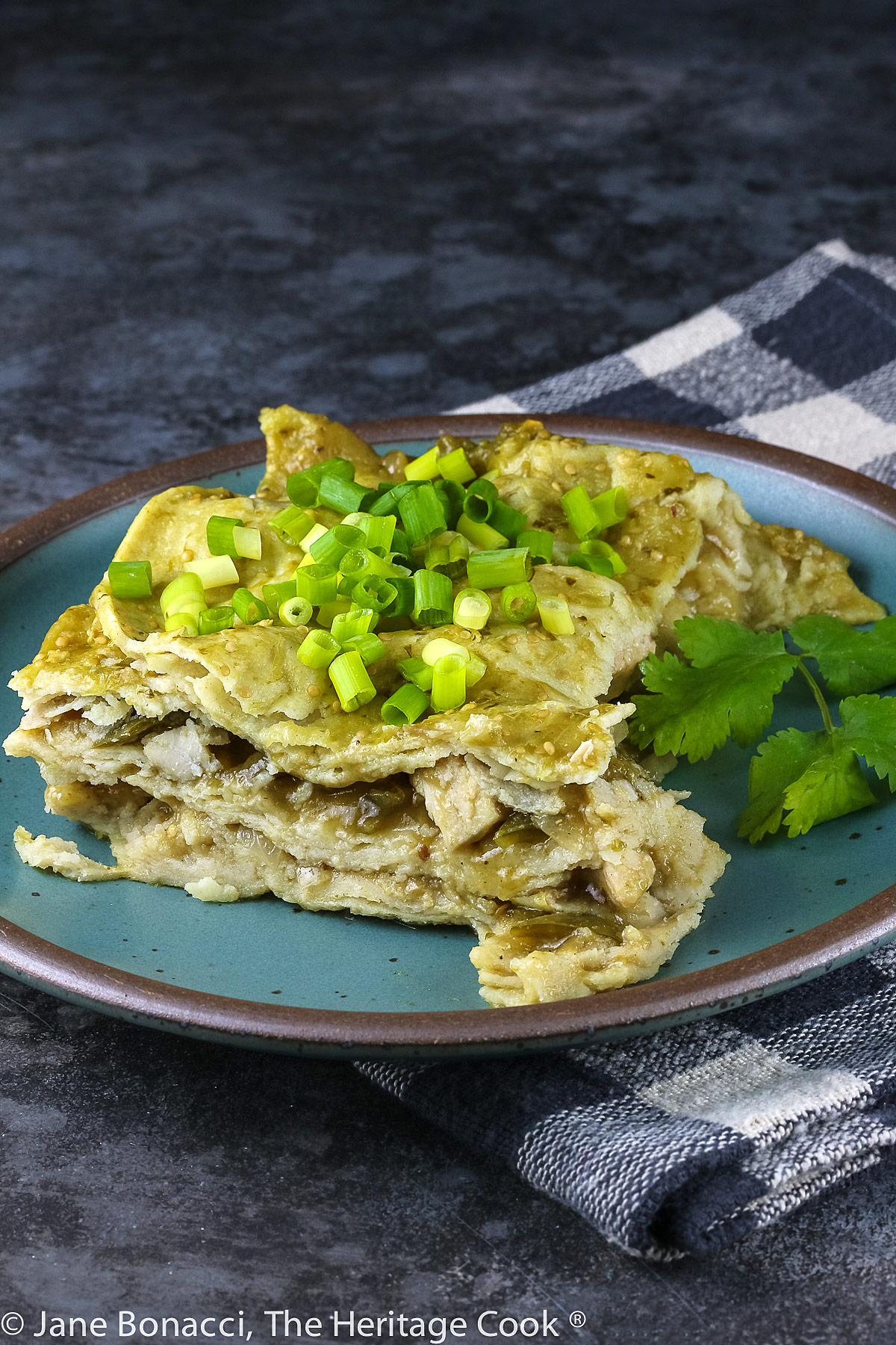 Square slice of the casserole on a teal colored plate with chopped green onions on top; Turkey Enchilada Casserole © 2022 Jane Bonacci, The Heritage Cook. 