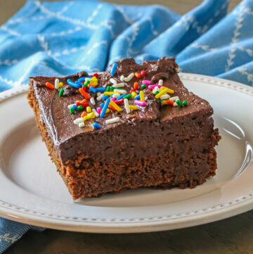 Square of Devils Food Chocolate Cake with dark chocolate frosting on a cream colored plate, some sprinkled with colorful sprinkles and a carafe of milk in the back © 2024 Jane Bonacci, The Heritage Cook.