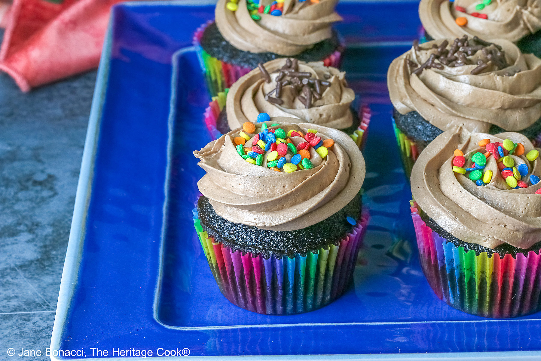 Chocolate cupcakes in rainbow colored paper liners with generously piped Nutella frosting and festive sprinkles on top, sitting on a blue rectangular platter with a pink cloth in the back; noAI © 2026 Jane Bonacci, The Heritage Cook.