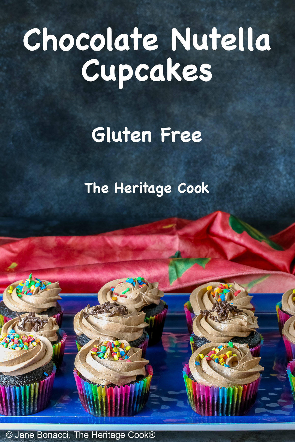 Chocolate cupcakes in rainbow colored paper liners with generously piped Nutella frosting and festive sprinkles on top, sitting on a blue rectangular platter with a pink cloth in the back; noAI © 2026 Jane Bonacci, The Heritage Cook.