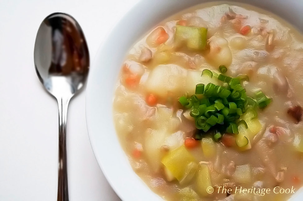 White bowl on white background filled with soup and topped with chopped chives or green onions; Corned Beef, Potato, and Leek Soup © 2023 Jane Bonacci, The Heritage Cook. 