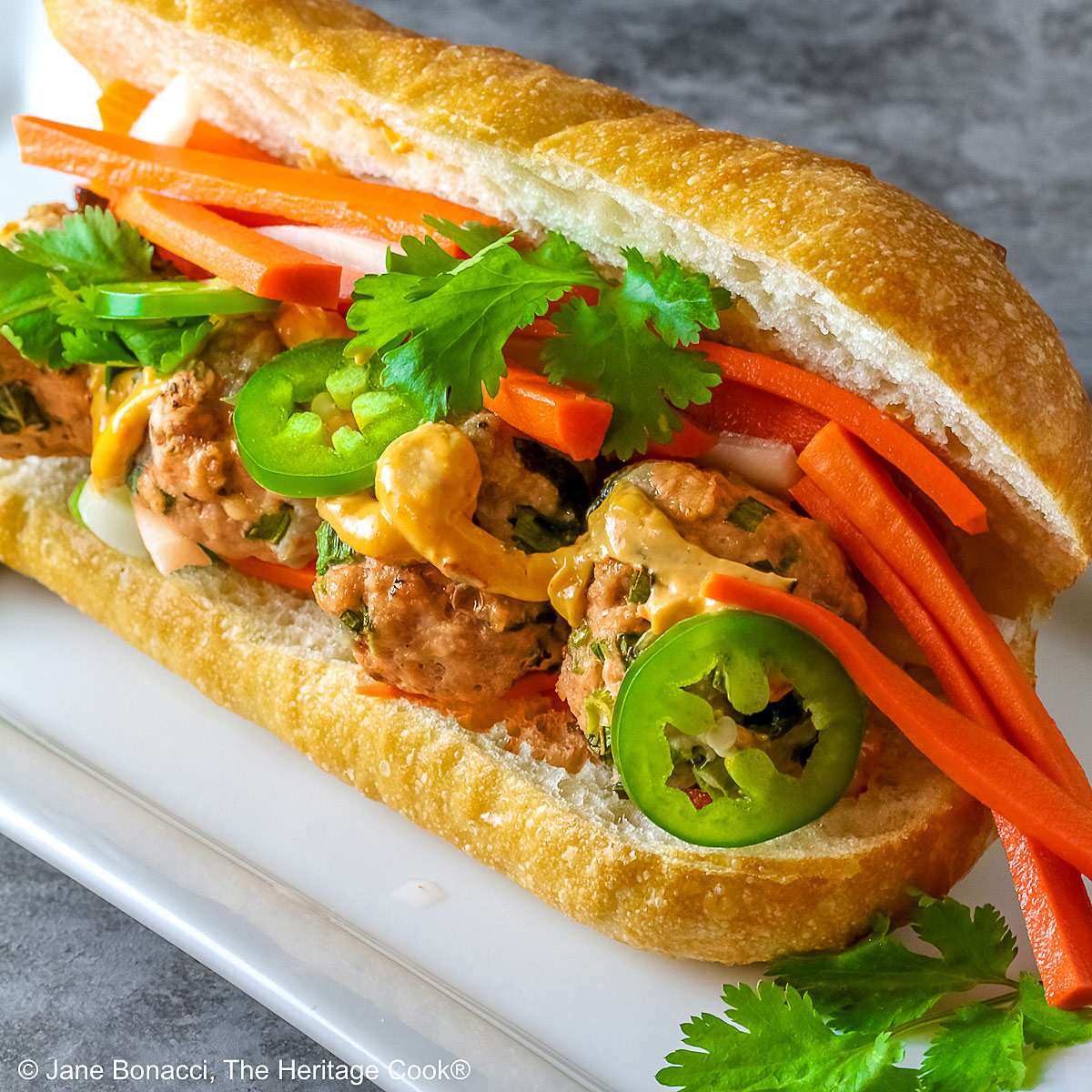 Brightly colored Banh Mi Pork Meatball Sandwich on a white rectangular plate filled with the meatballs, sliced green jalapeno peppers, pickled carrots and daikon radishes, cilantro leaves and a drizzle of the hot chili mayo with a jalapeno on one side © 2025 Jane Bonacci, The Heritage Cook.