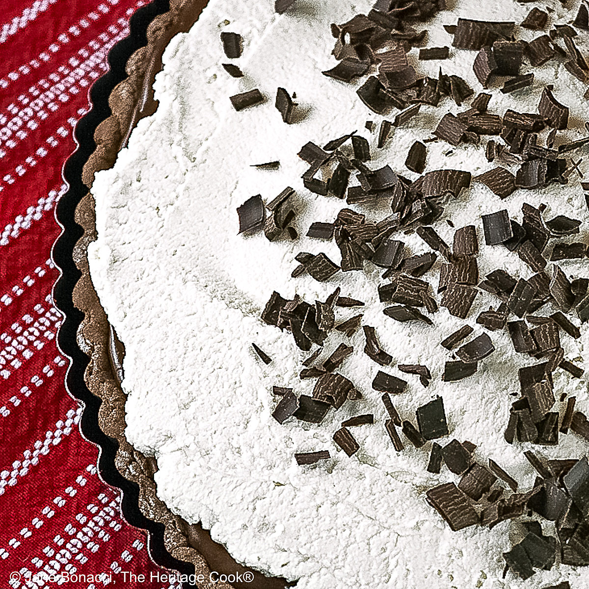 Chocolate Mocha Tart the whole tart, with the chocolate filling covered with a whipped topping and sprinkled with shaved chocolate pieces, some with a slice on a plate in front of the rest of the tart on red and white cloths © 2025 Jane Bonacci, The Heritage Cook.