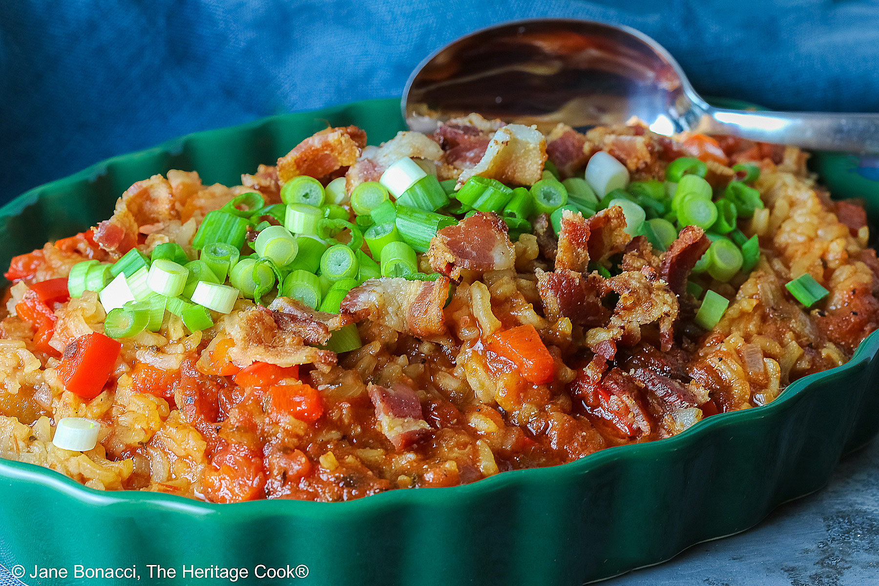 Shallow dark green au gratin dish filled until mounded with Spanish Rice with a Texas Twist topped with chopped green onions and bacon sitting on a blue cloth © 2025 Jane Bonacci, The Heritage Cook.