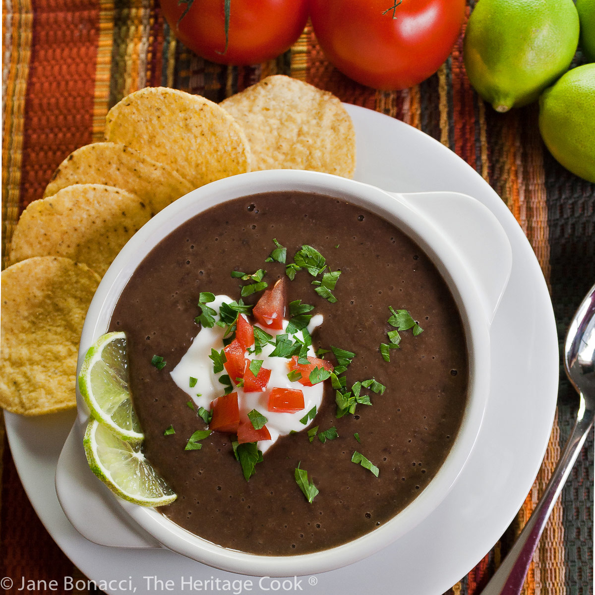Dark black bean soup in white bowls garnished with cream, tomatoes, and chopped cilantro and lime slices, with tortilla chips alongside; Chipotle Black Bean Soup with Cilantro Lime Cream (GF) © 2023 Jane Bonacci, The Heritage Cook.