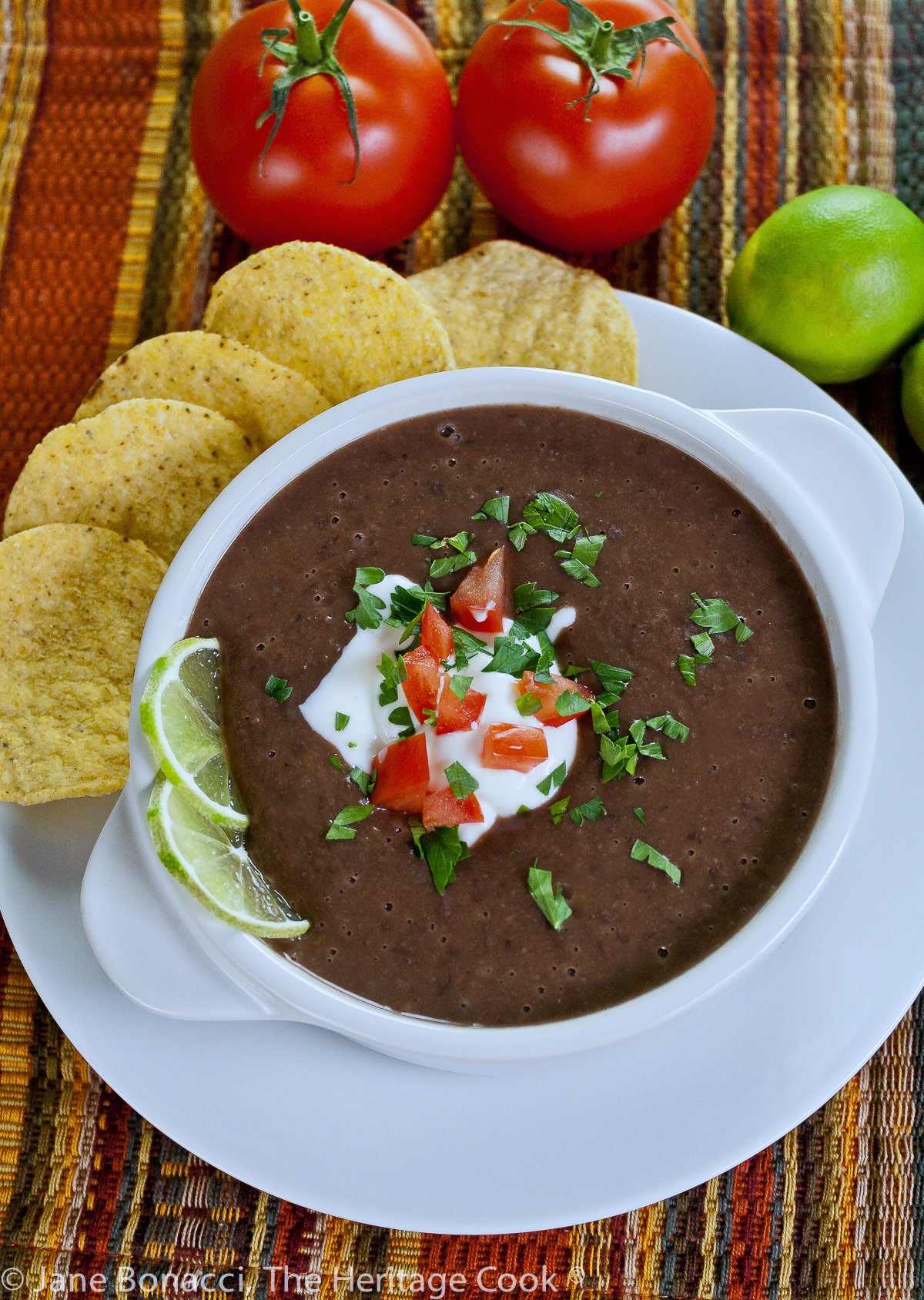 Dark black bean soup in white bowls garnished with cream, tomatoes, and chopped cilantro and lime slices, with tortilla chips alongside; Chipotle Black Bean Soup with Cilantro Lime Cream (GF) © 2023 Jane Bonacci, The Heritage Cook. 