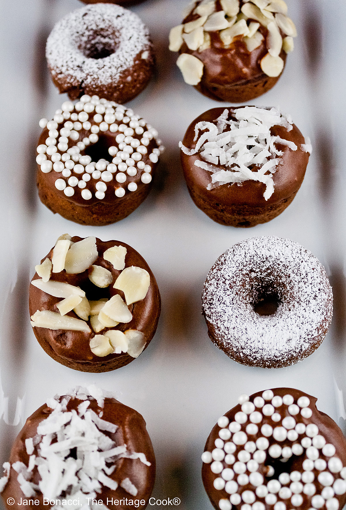 10 mini baked chocolate donuts topped with glaze and in a variety of white coverings, powdered sugar, coconut, almonds, etc. for a black and white presentation, sitting on a white rectangular platter with a vibrantly colored striped cloth beneath and a blue cloth behind © 2026 Jane Bonacci, The Heritage Cook.