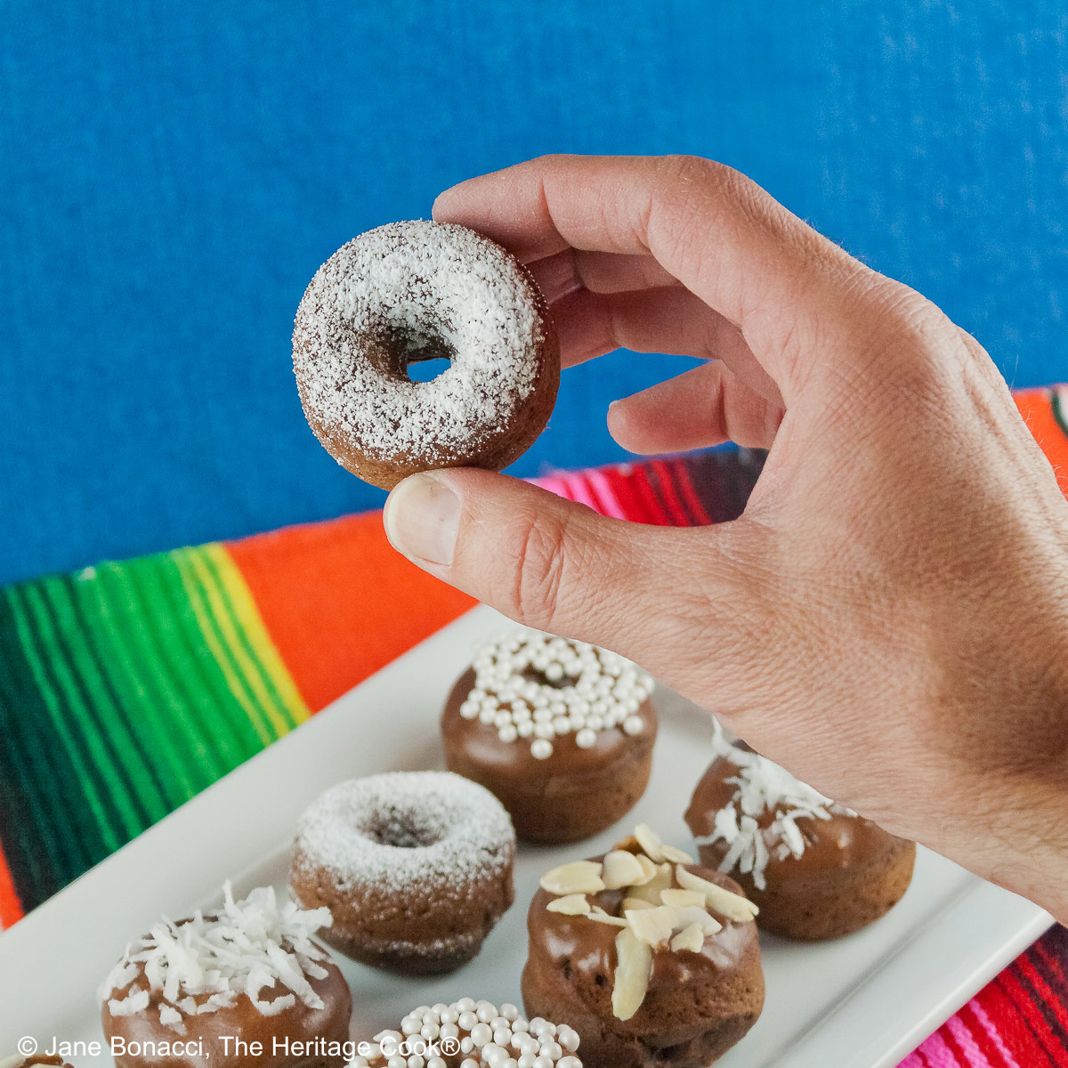 Gluten-Free Mini Baked Chocolate Cake Donuts copyright 2012 Jane Evans Bonacci, The Heritage Cook 10 mini baked chocolate donuts topped with glaze and in a variety of white coverings, powdered sugar, coconut, almonds, etc. for a black and white presentation, sitting on a white rectangular platter with a vibrantly colored striped cloth beneath and a blue cloth behind © 2026 Jane Bonacci, The Heritage Cook.
