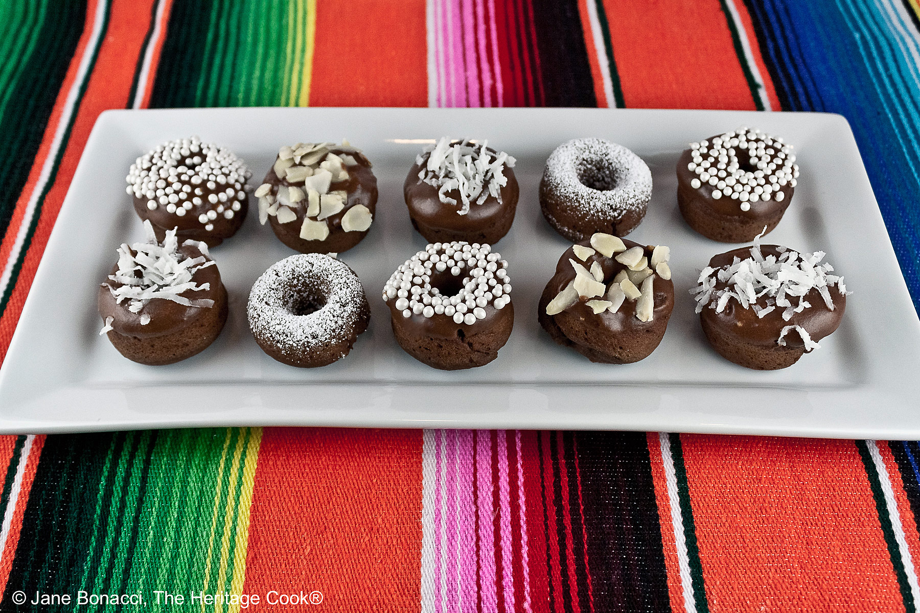Gluten-Free Mini Baked Chocolate Cake Donuts copyright 2012 Jane Evans Bonacci, The Heritage Cook 10 mini baked chocolate donuts topped with glaze and in a variety of white coverings, powdered sugar, coconut, almonds, etc. for a black and white presentation, sitting on a white rectangular platter with a vibrantly colored striped cloth beneath and a blue cloth behind © 2026 Jane Bonacci, The Heritage Cook.