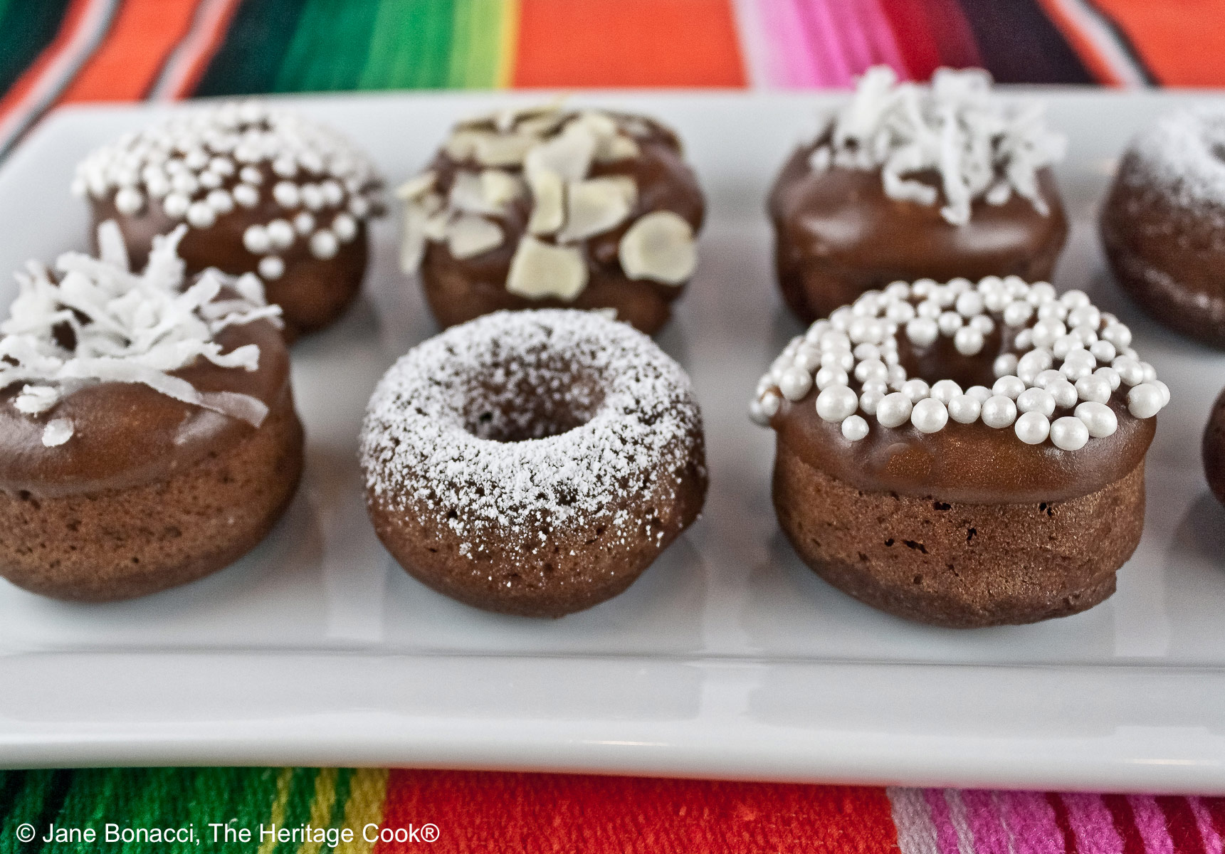 Gluten-Free Baked Mini Chocolate Cake Donuts copyright 2012 Jane Evans Bonacci, The Heritage Cook 10 mini baked chocolate donuts topped with glaze and in a variety of white coverings, powdered sugar, coconut, almonds, etc. for a black and white presentation, sitting on a white rectangular platter with a vibrantly colored striped cloth beneath and a blue cloth behind © 2026 Jane Bonacci, The Heritage Cook.