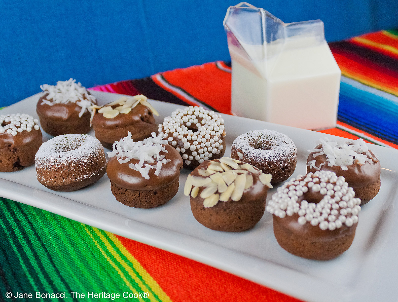 Gluten-Free Mini Baked Chocolate Cake Donuts copyright 2012 Jane Evans Bonacci, The Heritage Cook 10 mini baked chocolate donuts topped with glaze and in a variety of white coverings, powdered sugar, coconut, almonds, etc. for a black and white presentation, sitting on a white rectangular platter with a vibrantly colored striped cloth beneath and a blue cloth behind © 2026 Jane Bonacci, The Heritage Cook.