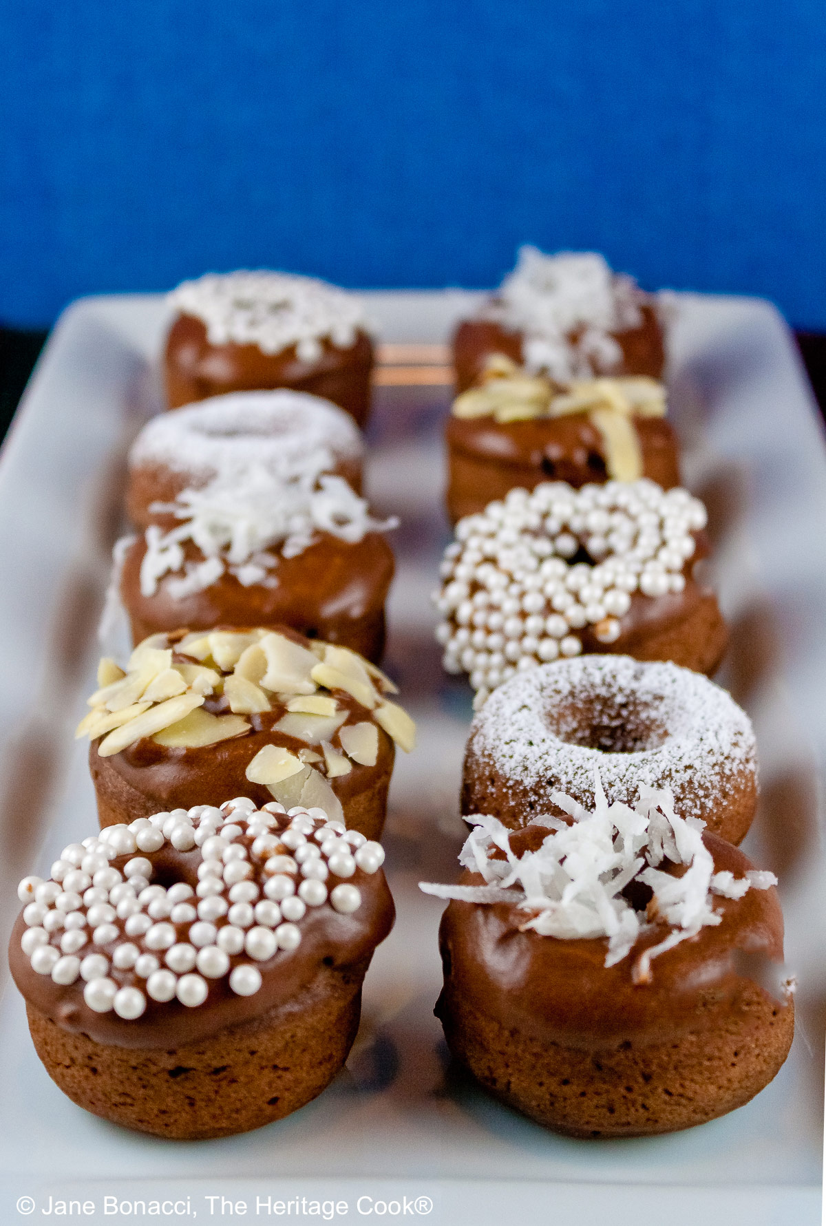 10 mini baked chocolate donuts topped with glaze and in a variety of white coverings, powdered sugar, coconut, almonds, etc. for a black and white presentation, sitting on a white rectangular platter with a vibrantly colored striped cloth beneath and a blue cloth behind © 2026 Jane Bonacci, The Heritage Cook.