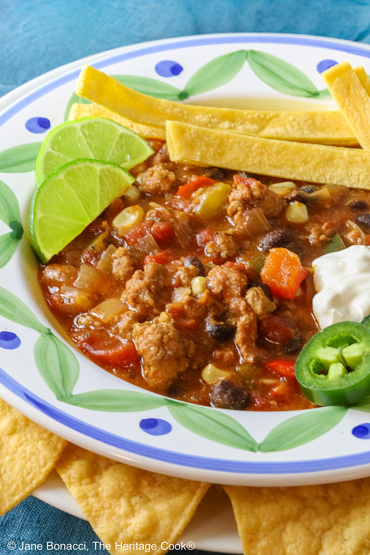 Pork and Chorizo Chili filling a bowl with a colorful decorated rim, sitting on a white plate, on top of an aqua cloth, with tortilla chips around the bowl; topped with lime slices, avocado slice, tortilla strips, sour cream, sliced jalapenos, chopped green onions © 2026 Jane Bonacci, The Heritage Cook.