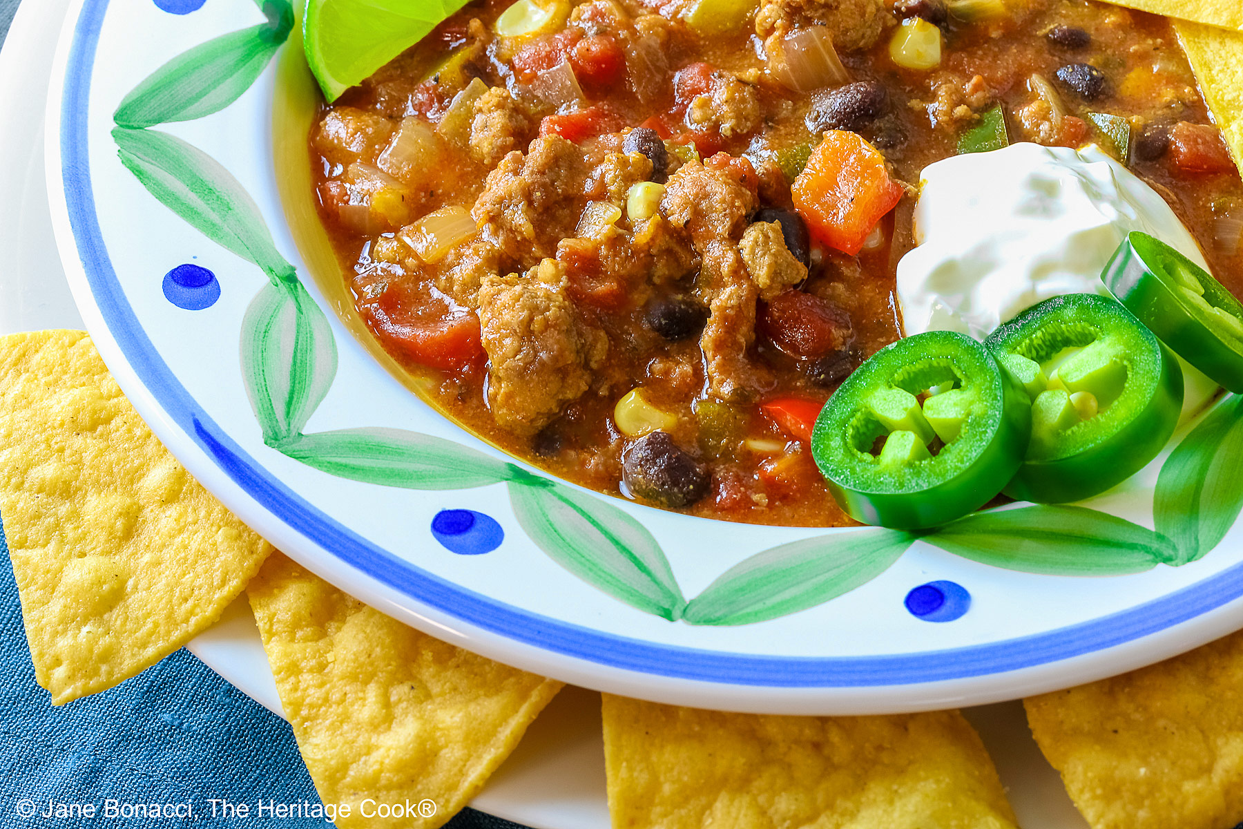 Del Monte Chili copyright 2012 Jane Evans Bonacci, The Heritage Cook Pork and Chorizo Chili filling a bowl with a colorful decorated rim, sitting on a white plate, on top of an aqua cloth, with tortilla chips around the bowl; topped with lime slices, avocado slice, tortilla strips, sour cream, sliced jalapenos, chopped green onions © 2026 Jane Bonacci, The Heritage Cook.