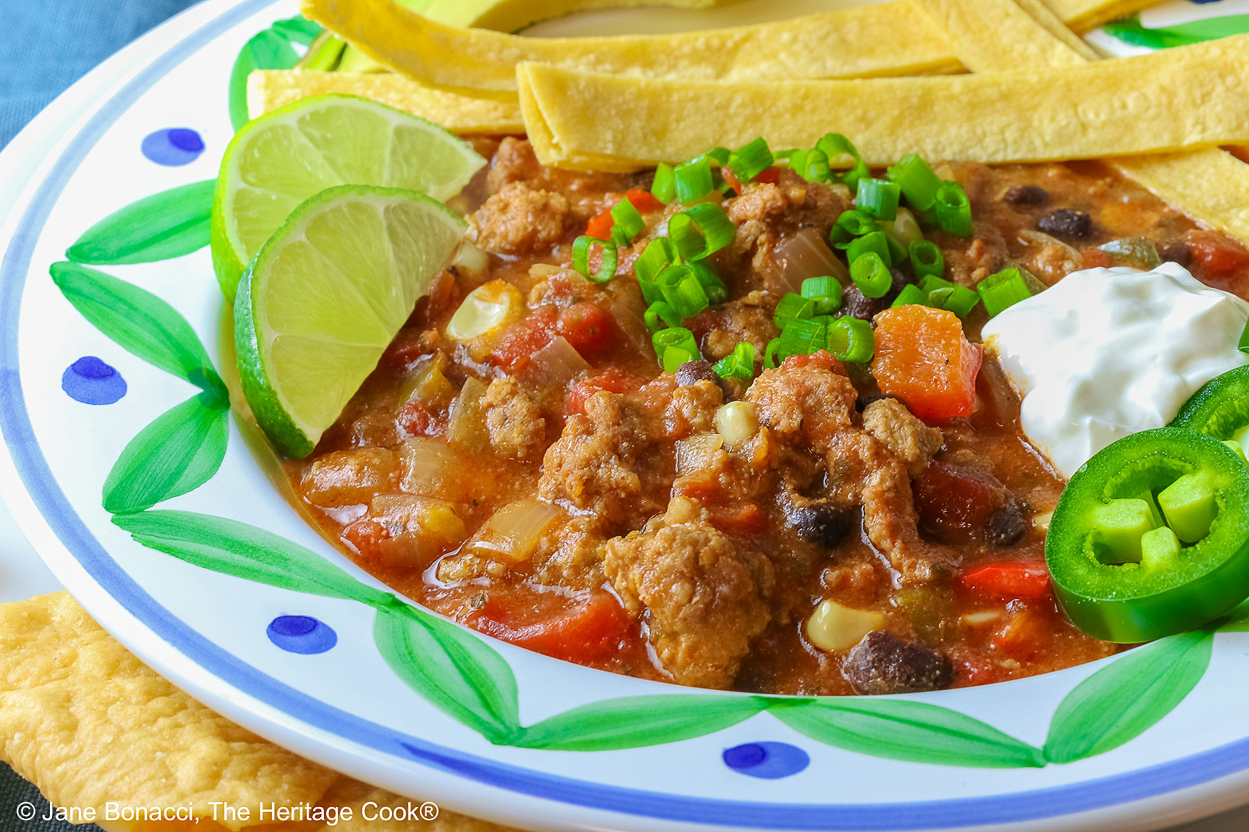Del Monte Chili copyright 2012 Jane Evans Bonacci, The Heritage Cook Pork and Chorizo Chili filling a bowl with a colorful decorated rim, sitting on a white plate, on top of an aqua cloth, with tortilla chips around the bowl; topped with lime slices, avocado slice, tortilla strips, sour cream, sliced jalapenos, chopped green onions © 2026 Jane Bonacci, The Heritage Cook.