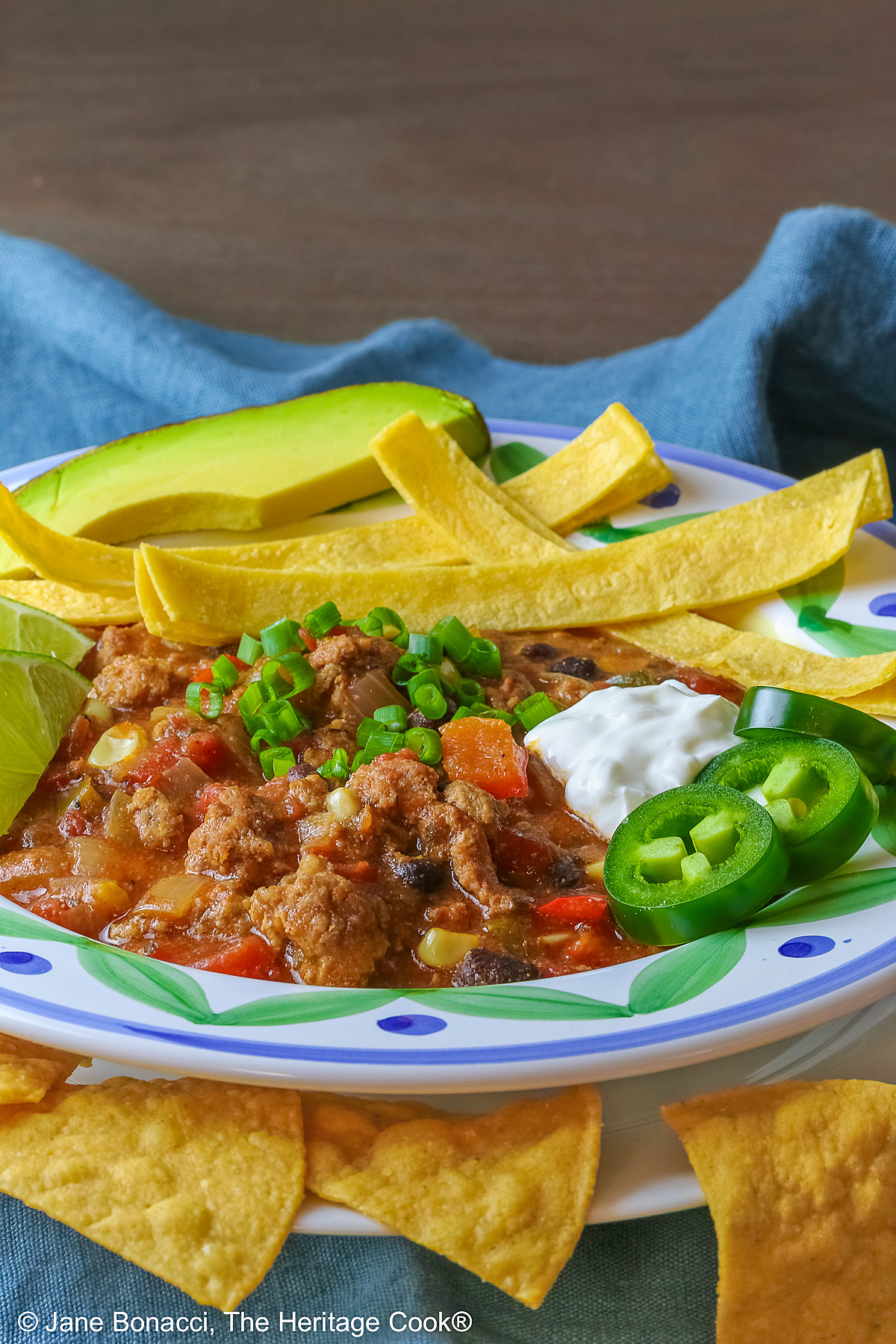 Del Monte Chili copyright 2012 Jane Evans Bonacci, The Heritage Cook Pork and Chorizo Chili filling a bowl with a colorful decorated rim, sitting on a white plate, on top of an aqua cloth, with tortilla chips around the bowl; topped with lime slices, avocado slice, tortilla strips, sour cream, sliced jalapenos, chopped green onions © 2026 Jane Bonacci, The Heritage Cook.