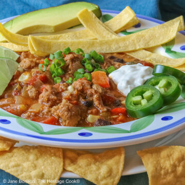 Pork and Chorizo Chili filling a bowl with a colorful decorated rim, sitting on a white plate, on top of an aqua cloth, with tortilla chips around the bowl; topped with lime slices, avocado slice, tortilla strips, sour cream, sliced jalapenos, chopped green onions © 2026 Jane Bonacci, The Heritage Cook.