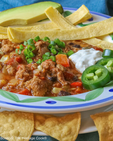 Pork and Chorizo Chili filling a bowl with a colorful decorated rim, sitting on a white plate, on top of an aqua cloth, with tortilla chips around the bowl; topped with lime slices, avocado slice, tortilla strips, sour cream, sliced jalapenos, chopped green onions © 2026 Jane Bonacci, The Heritage Cook.