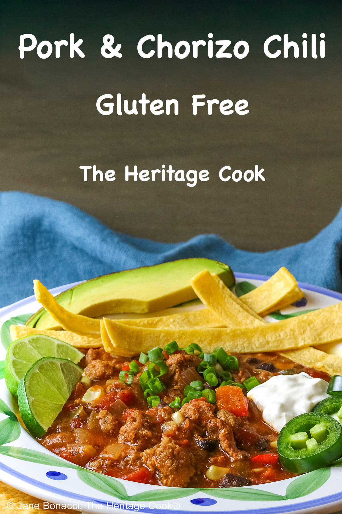 Pork and Chorizo Chili filling a bowl with a colorful decorated rim, sitting on a white plate, on top of an aqua cloth, with tortilla chips around the bowl; topped with lime slices, avocado slice, tortilla strips, sour cream, sliced jalapenos, chopped green onions © 2026 Jane Bonacci, The Heritage Cook.