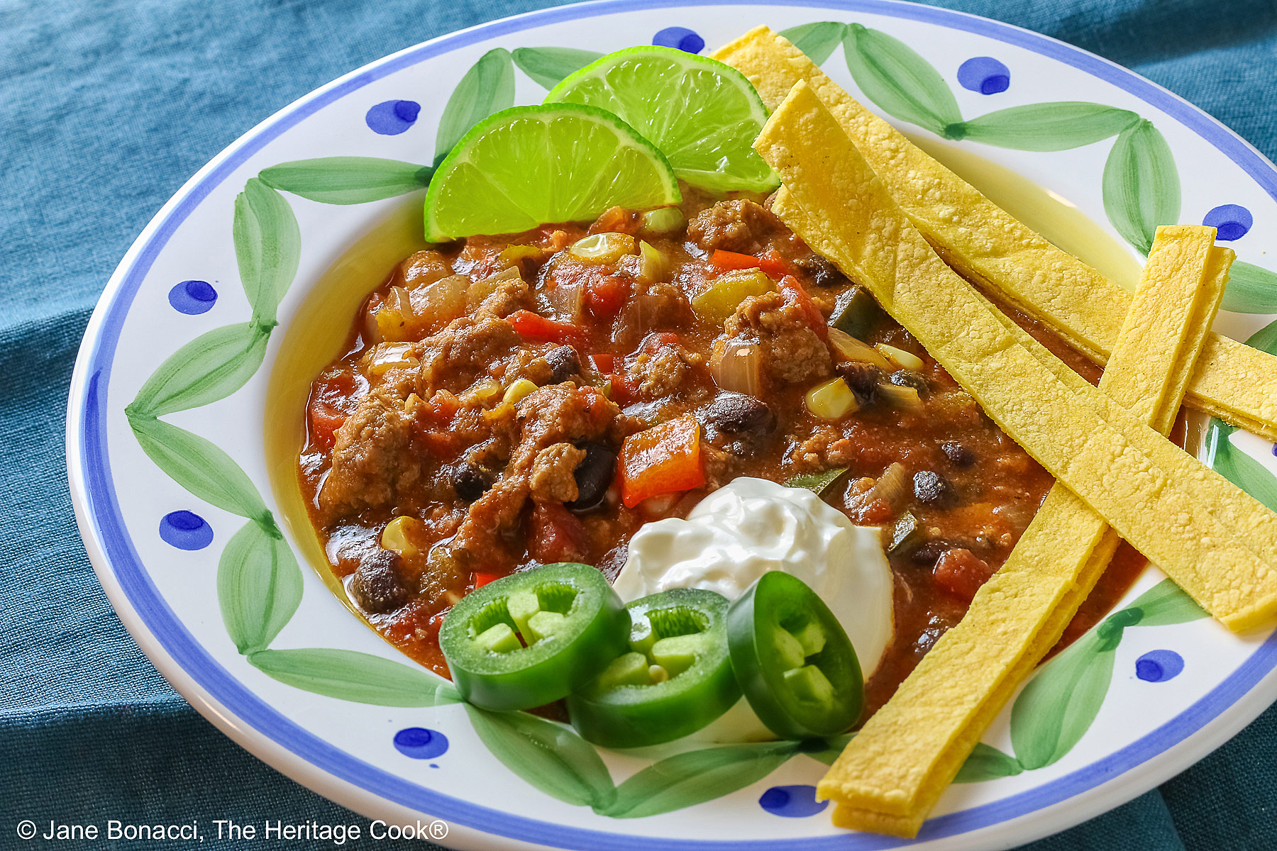 Pork and Chorizo Chili filling a bowl with a colorful decorated rim, sitting on a white plate, on top of an aqua cloth, with tortilla chips around the bowl; topped with lime slices, avocado slice, tortilla strips, sour cream, sliced jalapenos, chopped green onions © 2026 Jane Bonacci, The Heritage Cook.