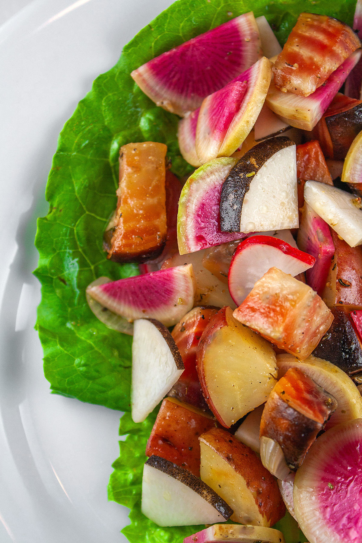 A serving of colorful cubed roasted potatoes and beets with crispy sliced radishes on a bright green lettuce leaf set on a white plate with a deep blue backdrop © Jane Bonacci, The Heritage Cook. 