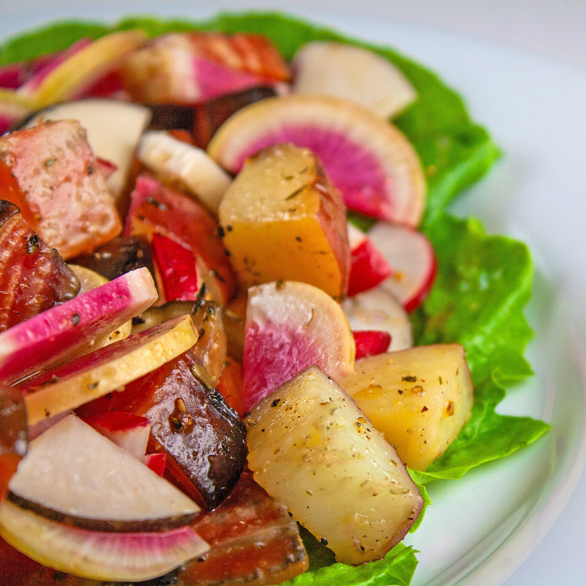 A serving of colorful cubed roasted potatoes and beets with crispy sliced radishes on a bright green lettuce leaf set on a white plate with a deep blue backdrop © Jane Bonacci, The Heritage Cook.