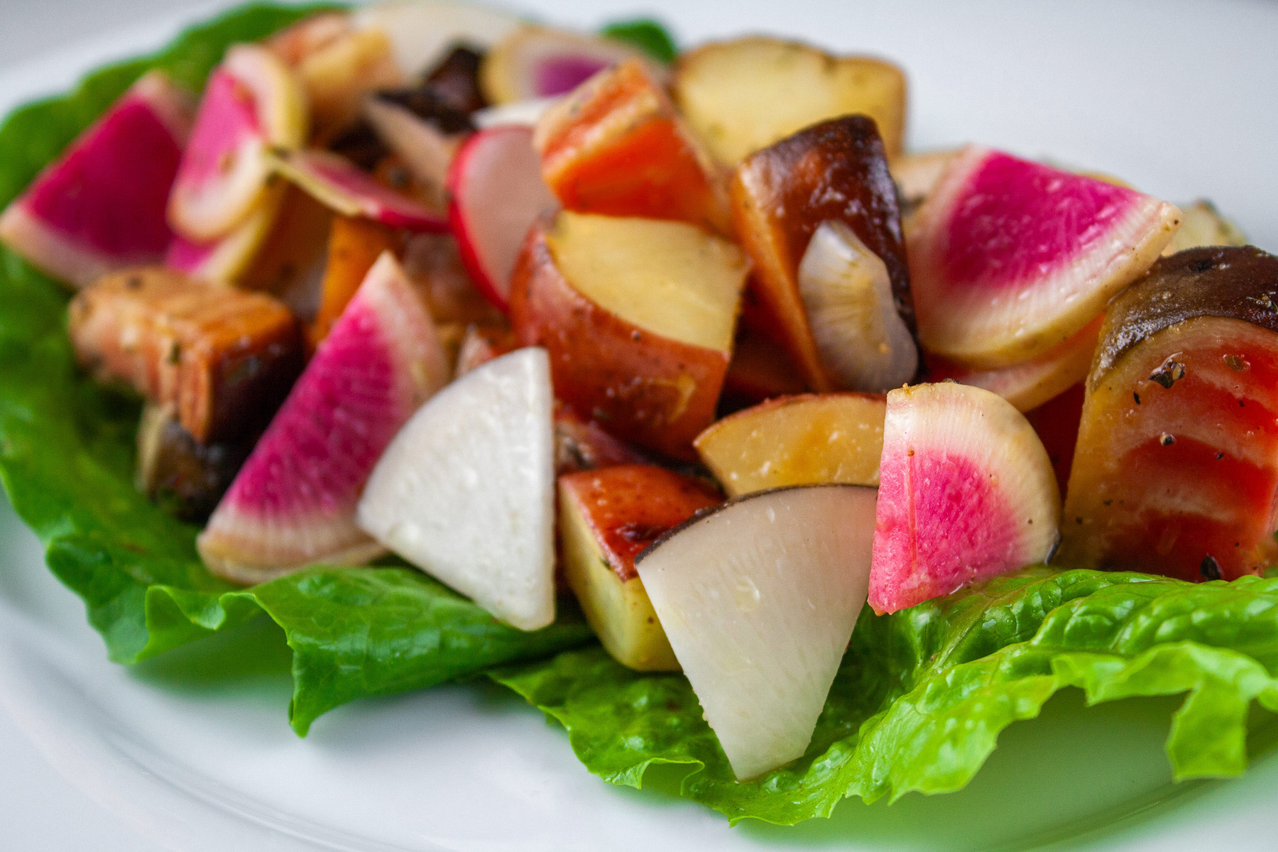 A serving of colorful cubed roasted potatoes and beets with crispy sliced radishes on a bright green lettuce leaf set on a white plate with a deep blue backdrop © Jane Bonacci, The Heritage Cook. 