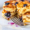 Stack of three blueberry pancakes on white plate with blue background; Blueberry Buttermilk Pancakes © 2023 Jane Bonacci, The Heritage Cook.
