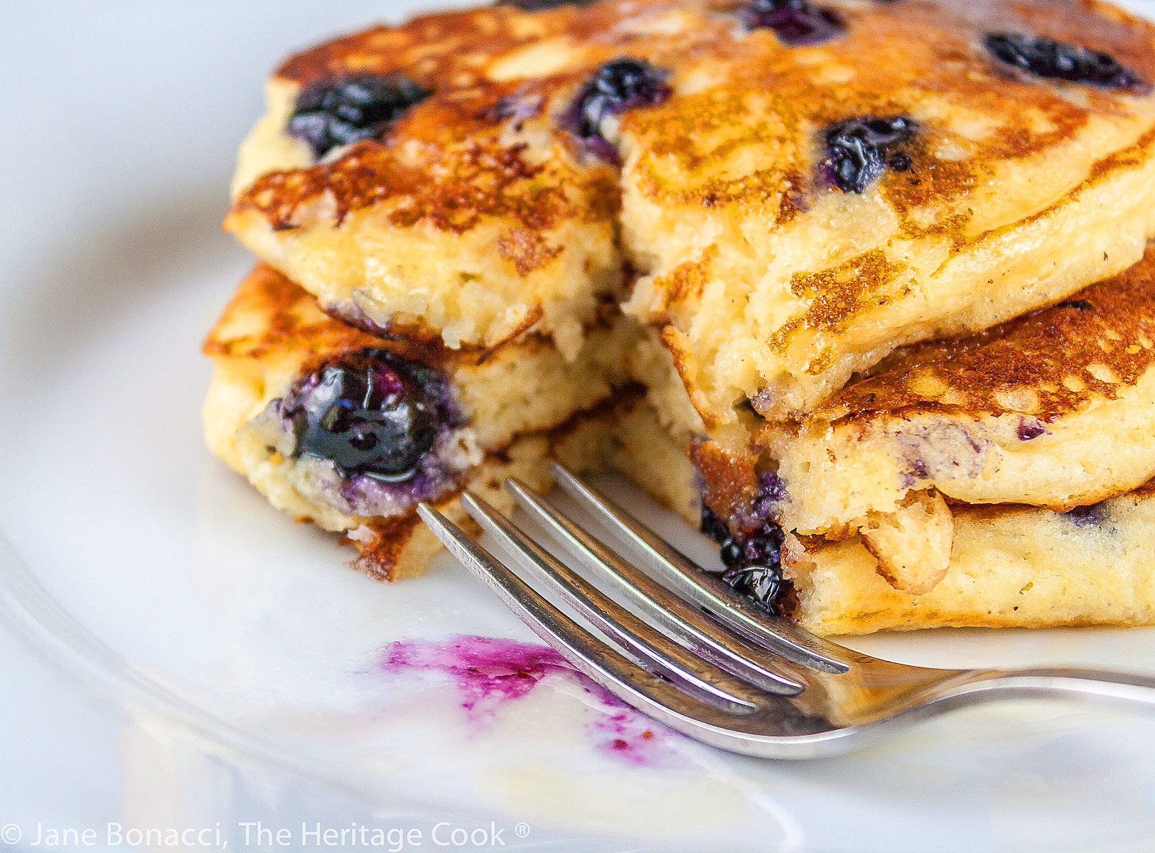 Stack of three blueberry pancakes on white plate with blue background; Blueberry Buttermilk Pancakes © 2023 Jane Bonacci, The Heritage Cook. 