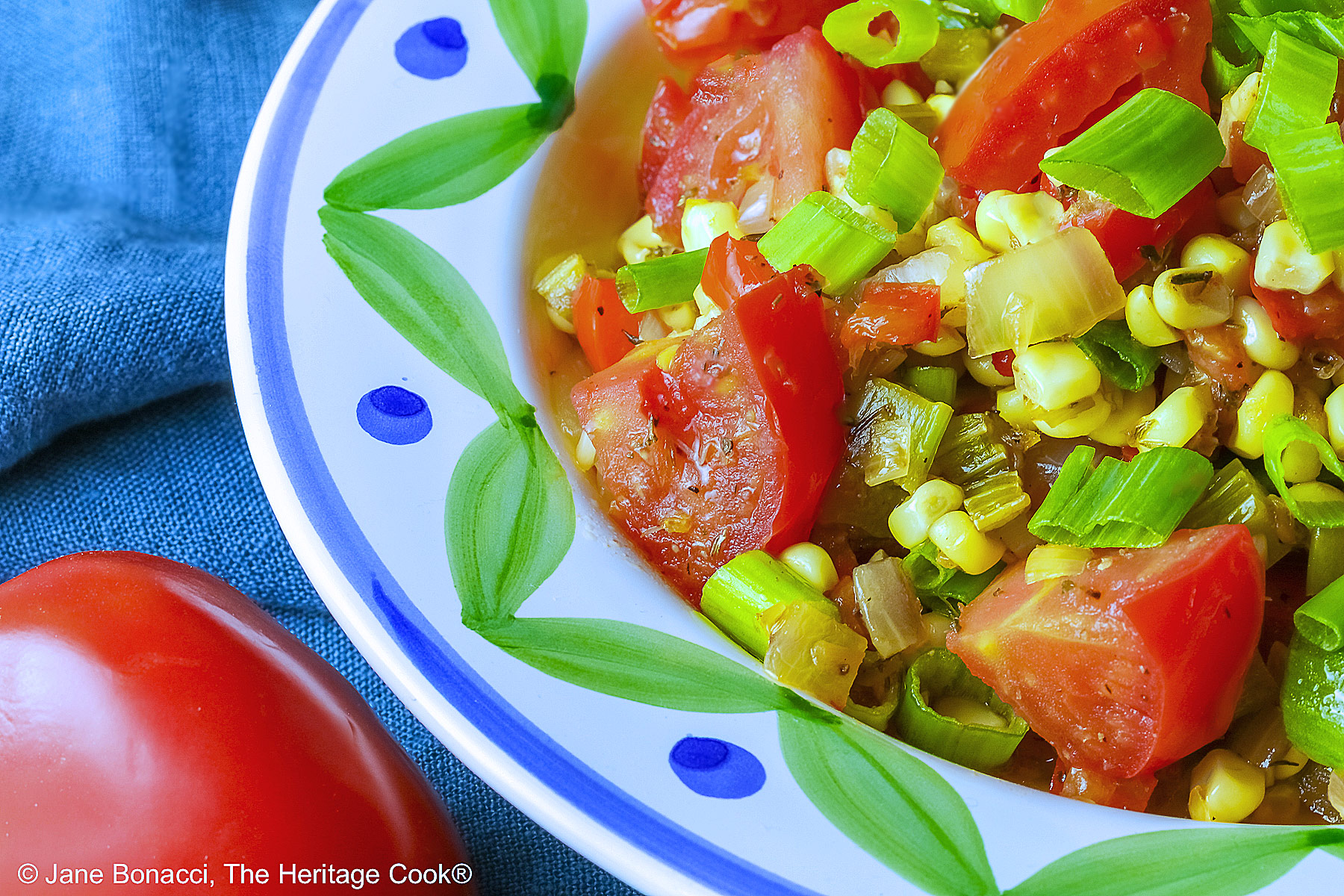 A bowl with a blue and green decorated rim filled with the yellow and red Fresh Corn and Tomato Salad with a piece of corn from the cob on top and a whole tomato in front sitting on a vivid blue cloth © 2026 Jane Bonacci, The Heritage Cook.