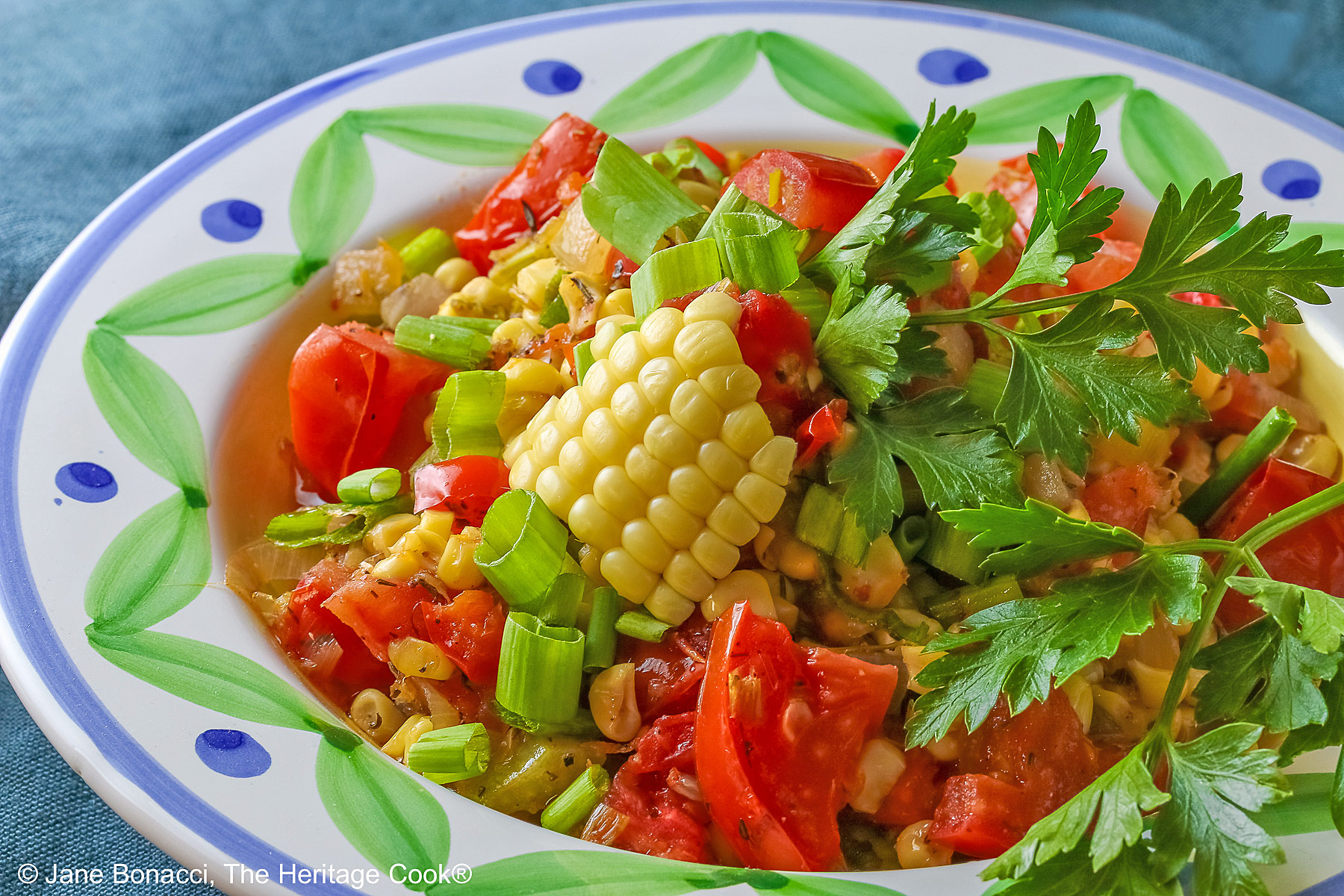 A bowl with a blue and green decorated rim filled with the yellow and red Fresh Corn and Tomato Salad with a piece of corn from the cob on top and a whole tomato in front sitting on a vivid blue cloth © 2026 Jane Bonacci, The Heritage Cook.