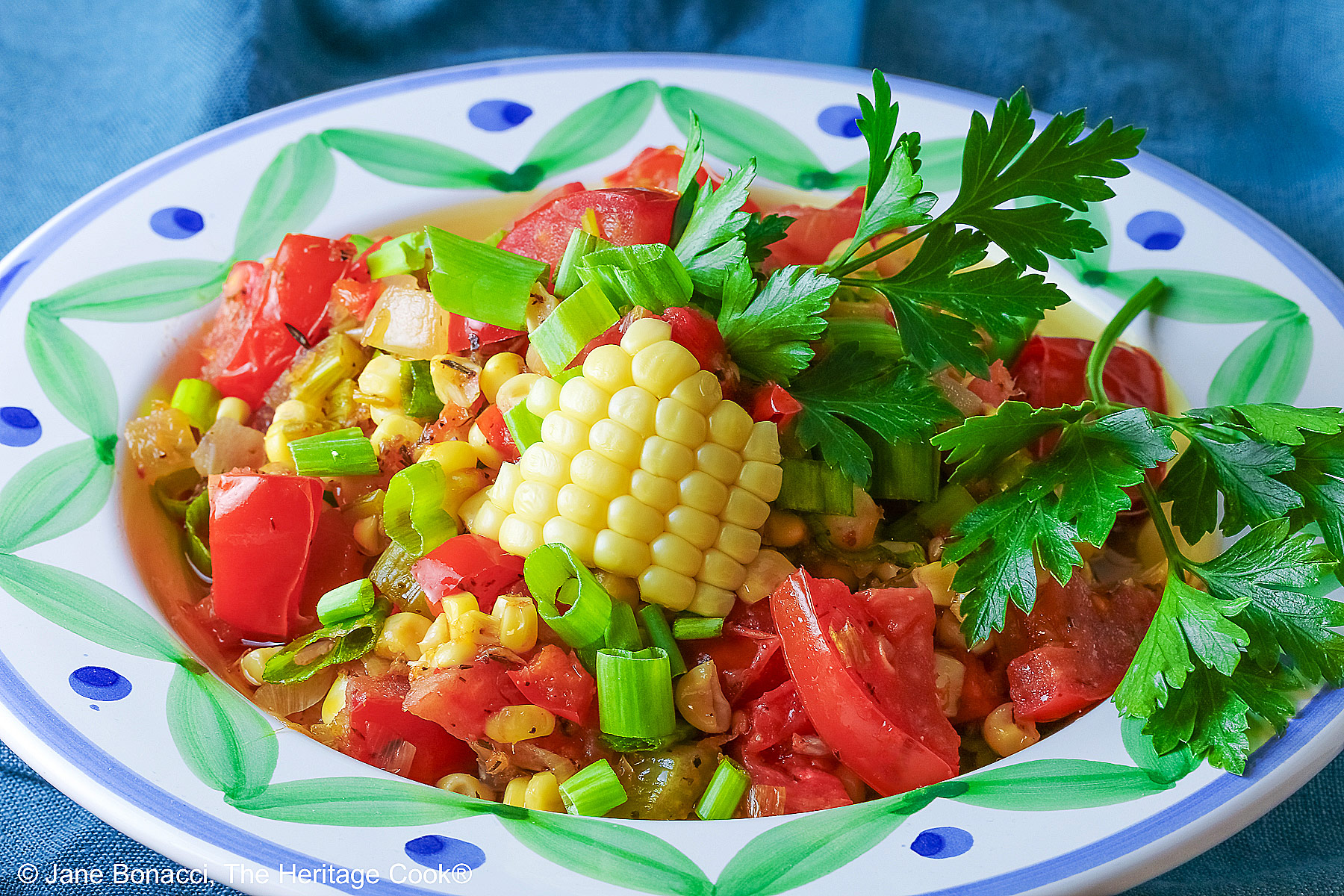 A bowl with a blue and green decorated rim filled with the yellow and red Fresh Corn and Tomato Salad with a piece of corn from the cob on top and a whole tomato in front sitting on a vivid blue cloth © 2026 Jane Bonacci, The Heritage Cook.