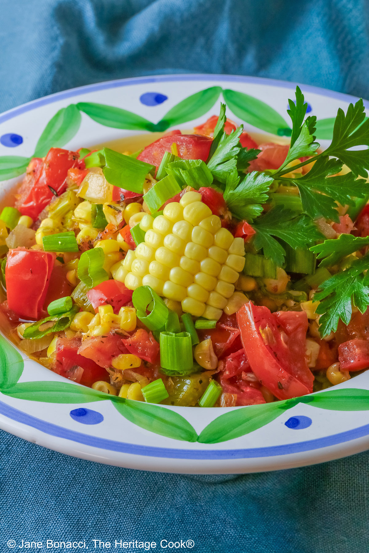 A bowl with a blue and green decorated rim filled with the yellow and red Fresh Corn and Tomato Salad with a piece of corn from the cob on top and a whole tomato in front sitting on a vivid blue cloth © 2026 Jane Bonacci, The Heritage Cook.
